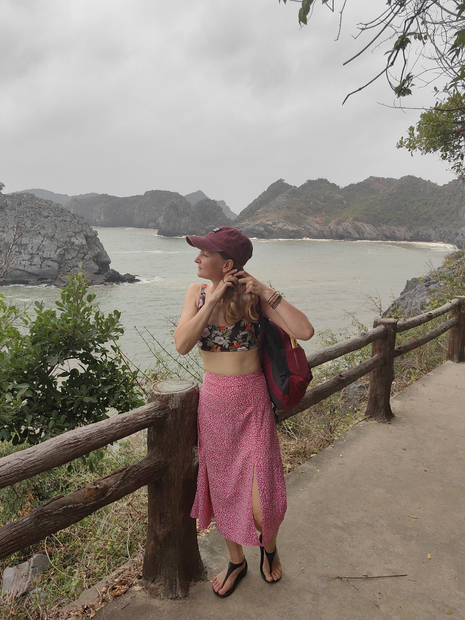 A woman in a floral bikini top, pink skirt with a slit, and a maroon baseball cap stands on a concrete path with a wooden railing, overlooking a body of water with rocky islands. She has a maroon backpack on one shoulder and is adjusting her hair. The sky is overcast, suggesting a cloudy or possibly rainy day. The location is likely in the city of Hải Phòng, Vietnam, given the tropical vegetation and coastal landscape.