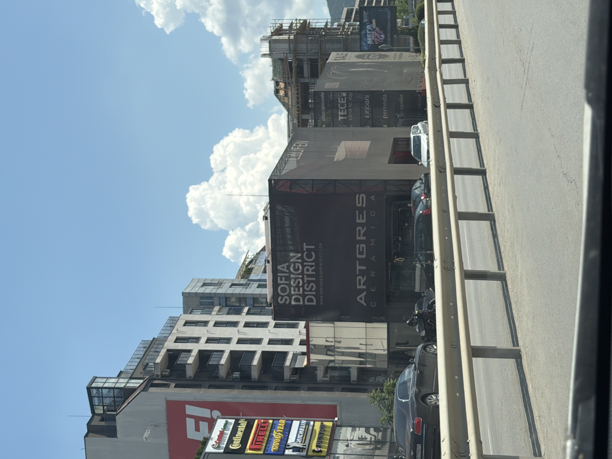 The image depicts a street scene in Sofia, Bulgaria, on a sunny day with scattered clouds. In the foreground, a metal guardrail runs along a road. Behind the guardrail, several cars are parked or driving, including a dark-colored Bentley and a white hatchback. A motorcycle with a rider is also visible.
Dominating the background are modern buildings, some of which appear to be under construction. Several large billboards and building facades display advertisements. Notable brands and businesses advertised include "Continental," "Pirelli," "Goodyear," "Michelin," "Dunlop," "Sofia Design District," "Artgres Ceramica," and "Laufen." There are also signs for "Tece," "Grohe," and "Sale" with a percentage discount. The overall impression is of a commercial and urban area in Sofia.