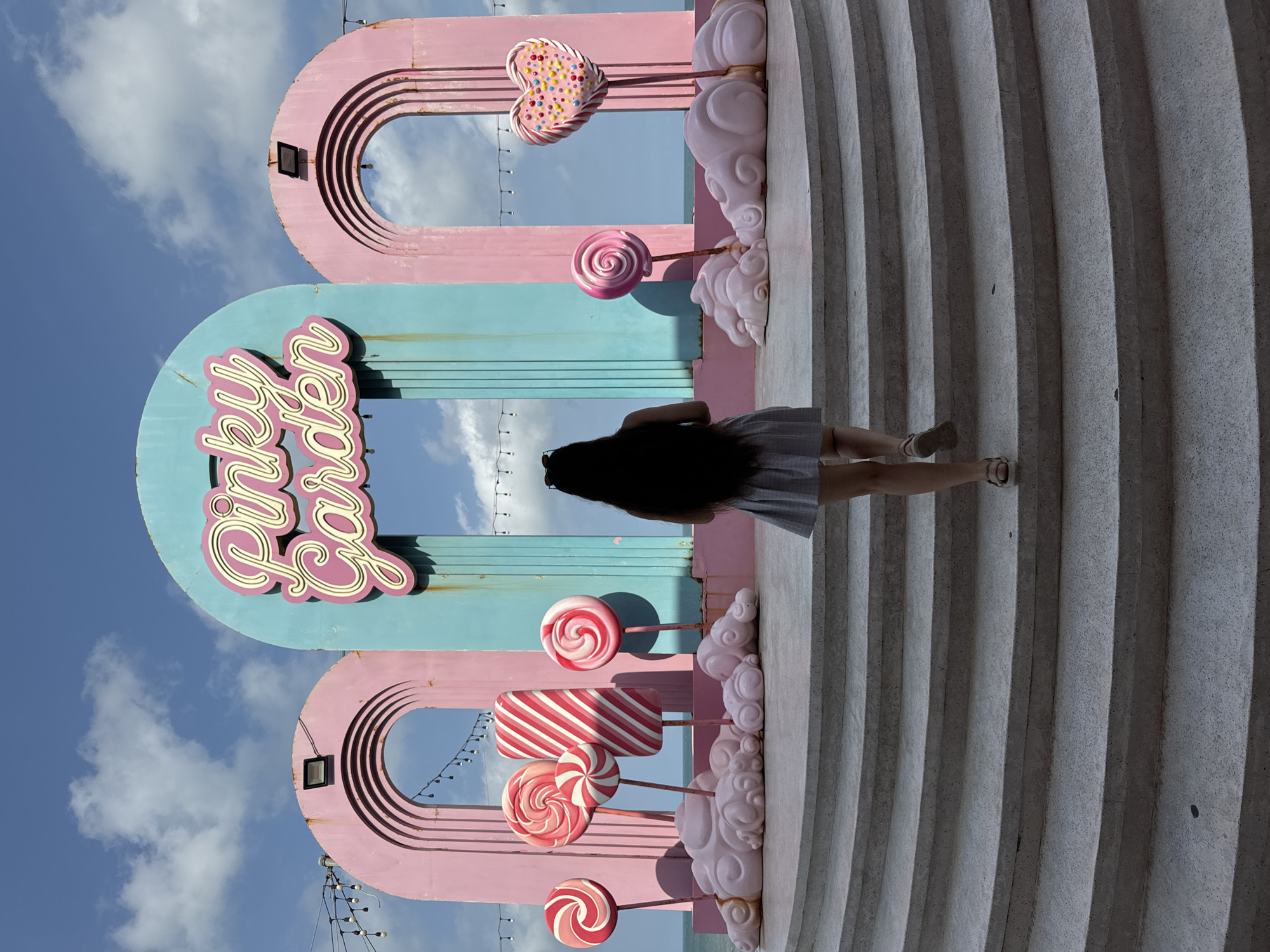 A woman with long dark hair, wearing a grey and white checkered skirt and sandals, walks up a set of wide, curving concrete stairs. She is walking towards a large, colorful installation featuring three arched structures. The central arch is turquoise and displays the neon sign "Pinky Garden" in pink script. The two flanking arches are pink.
In front of the arches, arranged on pink cloud-like structures, are several oversized lollipop and candy-shaped props. These include three round lollipops with red and white swirls, a rectangular candy with red and white diagonal stripes, a pink swirl lollipop, and a pink heart-shaped candy adorned with colorful sprinkles. Strings of lights hang across the tops of the arches.
The background shows a bright blue sky with scattered white clouds and the ocean. This installation is located in Tien Thanh Ward, Vietnam, a coastal area known for its beaches and tourist attractions. The overall scene suggests a whimsical and playful outdoor setting.