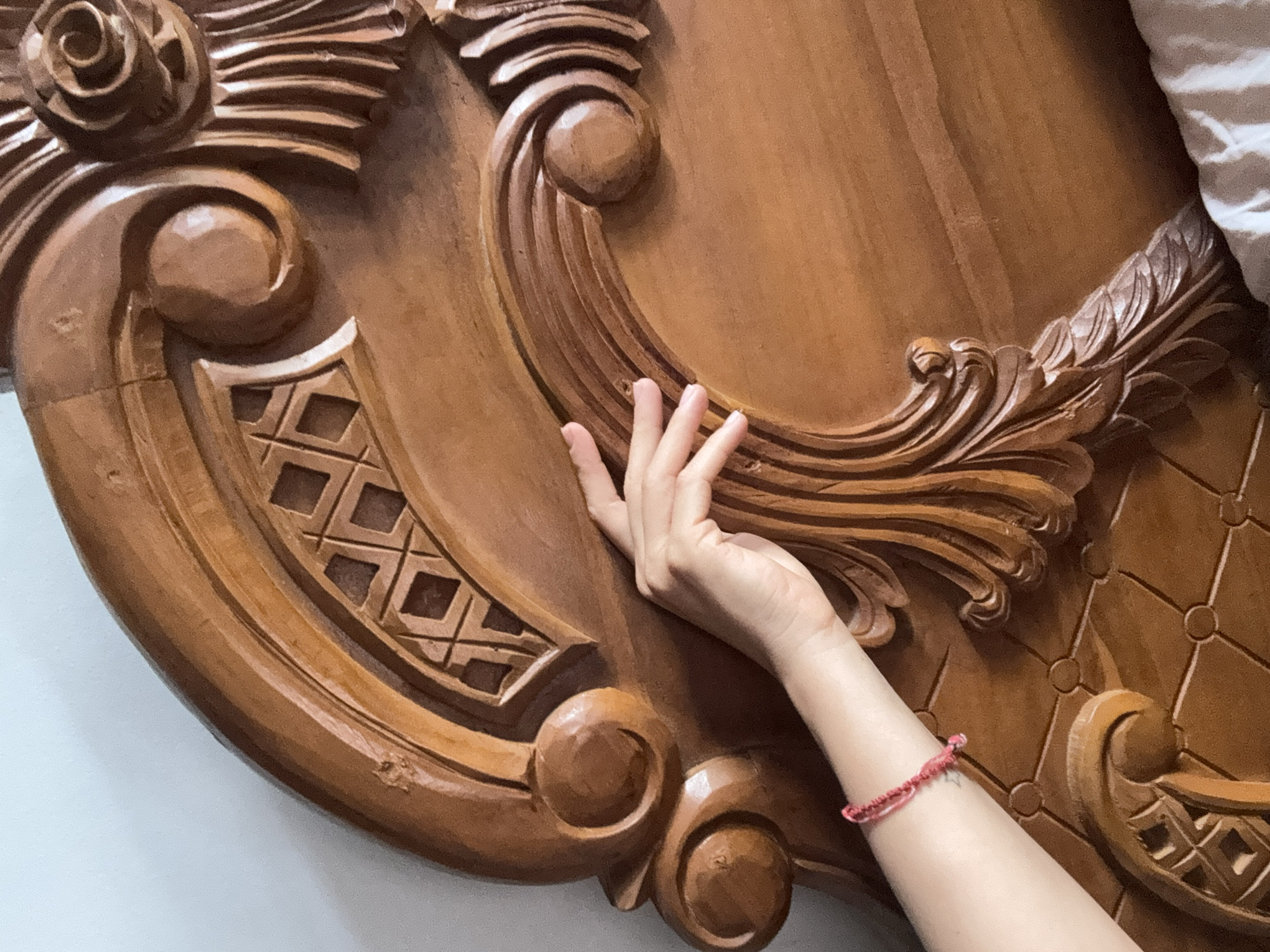 A close-up, eye-level indoor shot depicts a hand resting on the carved headboard of a wooden bed. The hand is light-skinned, with extended fingers. A red braided bracelet adorns the wrist. The headboard is ornate, featuring a variety of carved patterns. To the left, the wall is white. To the right, a white pillow is seen. The location is Kuta Selatan, Indonesia.