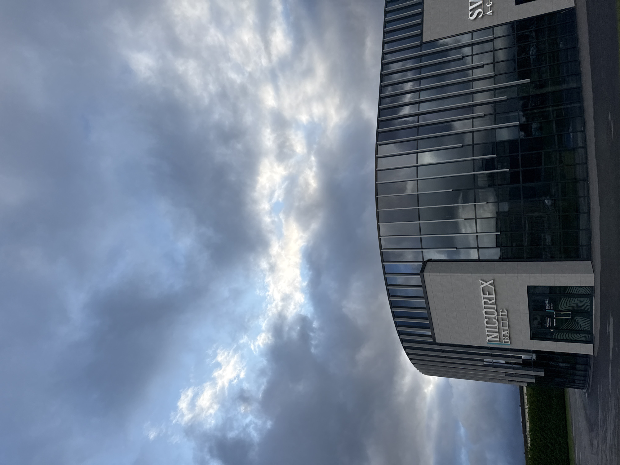 The image shows the Nicorex Baltic building, located in Peetri alevik, Estonia. The building features a modern, curved architectural design with a facade composed of grey panels and large, reflective glass windows. The Nicorex Baltic logo is prominently displayed in white lettering on the grey panel section of the building. An entrance with double glass doors is visible, also featuring signage. The sky above is dramatic, with a mix of dark storm clouds and patches of bright sky. To the left of the building, a dense green hedge forms a boundary, and a paved area or parking lot is in the foreground. The overall scene suggests a business or industrial complex under a moody sky.