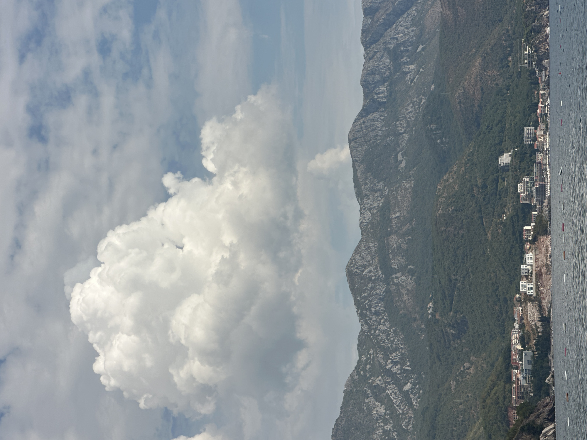 The image depicts a panoramic view of the coastline of Rafailovići, Montenegro. In the foreground, the calm, dark blue sea is visible, with a series of white and red buoys marking the water. The middle ground features a densely populated coastline with numerous buildings, ranging from modern white structures to older, multi-story complexes with terracotta roofs. These buildings are nestled along the shore at the base of a steep, green, mountainous terrain. The mountains are characterized by their rugged, rocky slopes and are covered in dense vegetation in the lower sections. In the sky above, large, dramatic white cumulus clouds dominate the scene, set against a backdrop of lighter blue sky with scattered wisps of clouds. The overall atmosphere is serene, with a clear division between the water, the populated coast, and the imposing natural landscape.