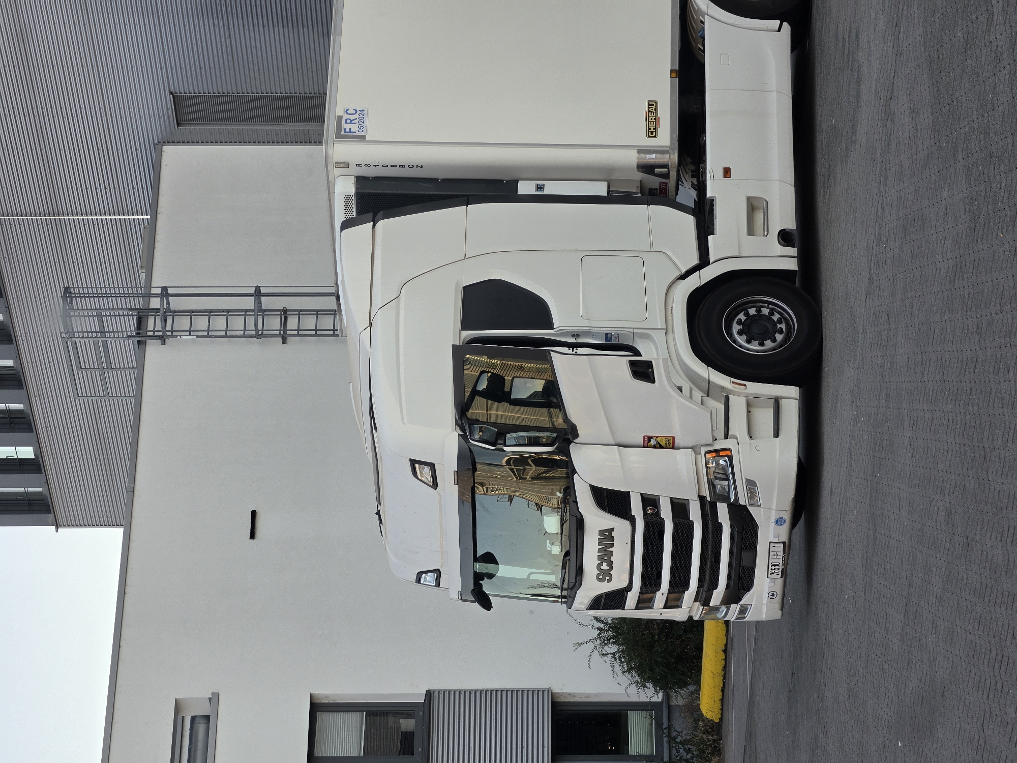 A white Scania semi-truck is parked in an industrial area. The truck has a white cab with black accents and a large white trailer with the "CHEREAU" logo. The trailer also has a blue label with "FRC 05/2024". The truck is parked on a gray paved surface in front of a modern white building with horizontal gray siding on the upper sections and a gray metal ladder on the exterior. The background features a portion of a tall building with horizontal gray corrugated metal siding and windows. The sky is overcast and a soft yellow concrete barrier is visible on the left side of the image. The license plate on the truck reads "76580 11". The scene appears to be an industrial zone within the city of Frankfurt, Germany, likely a logistics or distribution center.