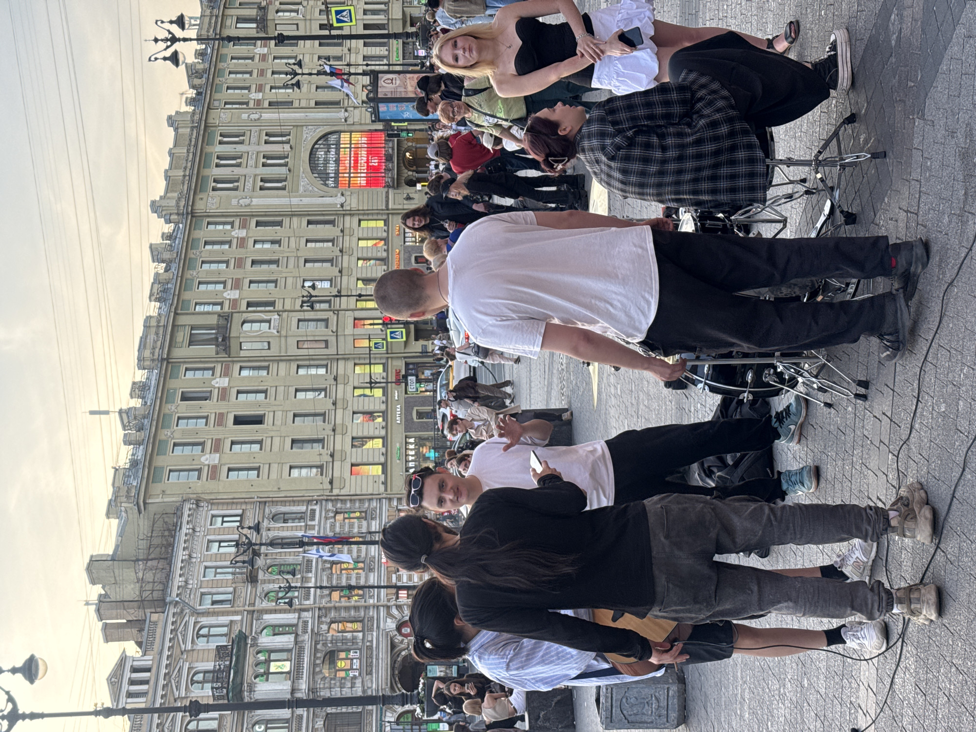 This media file depicts a street scene with a group of people gathered, likely for a performance or social event. The subjects include several individuals, some of whom appear to be musicians or performers. One person in the foreground is wearing a white t-shirt and black pants, facing away from the camera. Another individual in a black and white plaid shirt is seated, possibly playing drums, as drum equipment is visible. A blonde woman in a black top and white skirt is also present.
The setting is an urban environment with a grand, ornate building in the background, characterized by numerous windows and architectural details. The ground is paved with cobblestones. Street lamps and power lines are also visible, suggesting a city street.
The activity appears to be a street performance, with a gathering of onlookers. The time of day seems to be late afternoon or early evening, as indicated by the soft, warm lighting and the angle of the shadows. The weather appears to be clear.
Notable details include the diverse attire of the individuals, ranging from casual to more stylish outfits. The presence of a phone in one person's hand might suggest they are documenting the event or communicating.
There is visible text on a large illuminated sign on the building in the background, which appears to be in Cyrillic script. Some smaller signs with text are also present on the building's facade. The overall mood seems relaxed and engaging, with people observing and interacting.
