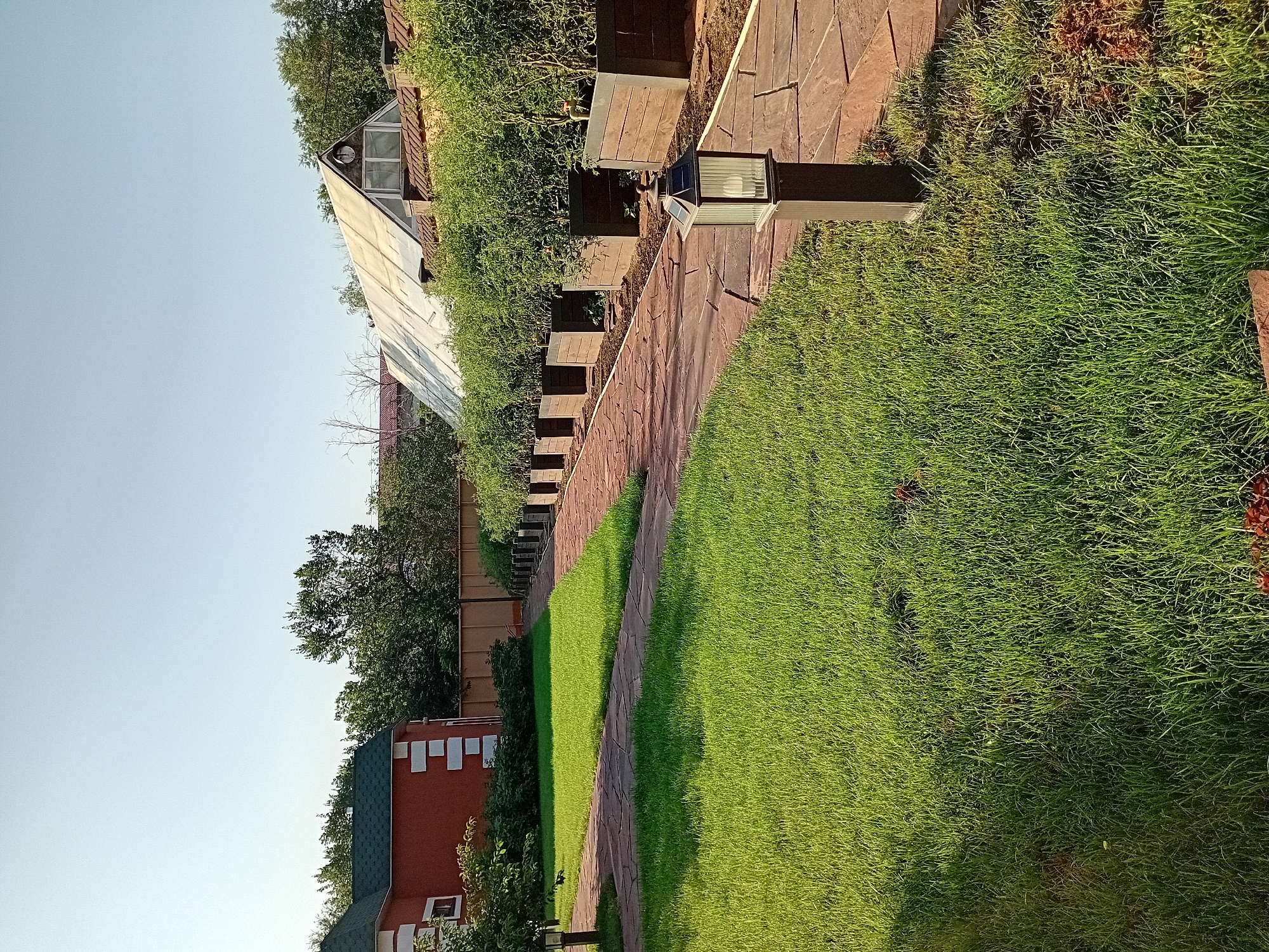 This is a full, eye-level, outdoor shot of a residential backyard in Semey, Kazakhstan, captured on a sunny day. A well-maintained, bright green lawn occupies the left side of the image, with a shadow cast from the left. A pathway made of large, reddish-brown flagstone tiles with a patterned surface winds from the foreground, curving to the right and disappearing into the background. Along the right side of the pathway, a row of dark wooden planter boxes filled with lush, green vegetation, likely tomato plants, are lined up. One black, rectangular garden lamp with a ribbed glass shade stands on a dark post along the pathway. In the background, a red brick house with a dark green roof and white trim is visible on the left. To the right of the house and behind the planters, a large, translucent, A-frame greenhouse with a white roof is partially visible. The sky is clear and pale blue.