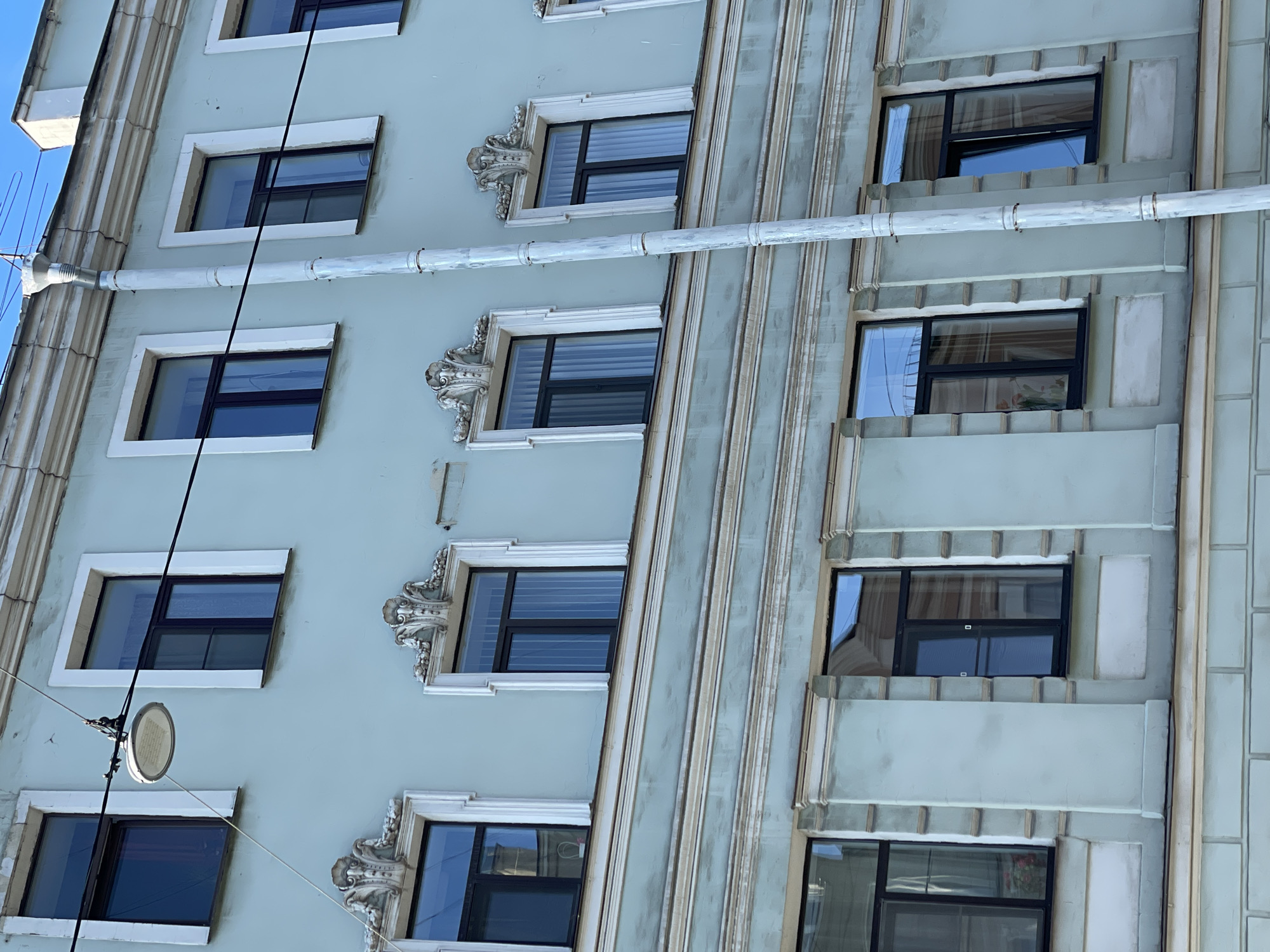 The image depicts the facade of a building, likely a multi-story residential or commercial structure, in Saint Petersburg, Russia. The architectural style appears to be historic, possibly dating back to the late 19th or early 20th century. The facade is characterized by a light-colored exterior (possibly light blue or grey) and features rows of windows with varying decorative surrounds. Some windows have elaborate ornate moldings above them, while others feature simpler, more geometric designs. There is a rain gutter or pipe running horizontally across the facade, intersecting with electrical cables. An exterior light fixture is visible on the left side of the building. The building's exterior details suggest that it is located in a historic district of Saint Petersburg, reflecting the city's rich architectural heritage.