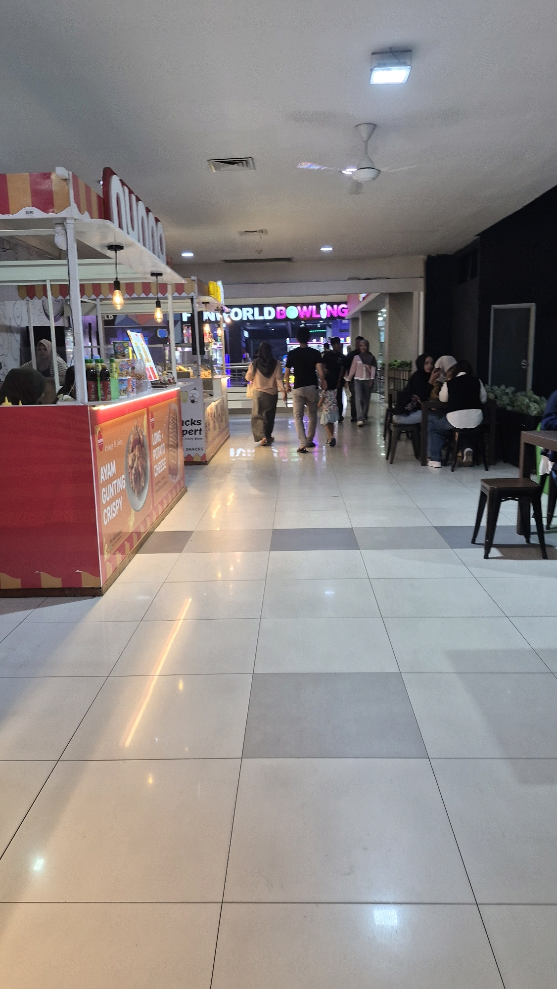 A nighttime indoor shot captures a view of a brightly lit shopping mall hallway with a polished tiled floor reflecting overhead lights. To the left, a food stall with a red facade displays signage for "AYAM GUNTING CRISPY" and "LONG POTATO CHEESE". Further down the hallway, a neon sign reads "WORLD BOWLING". Several individuals are walking in the hallway, some seated at tables. A ceiling fan is visible near the top of the frame.