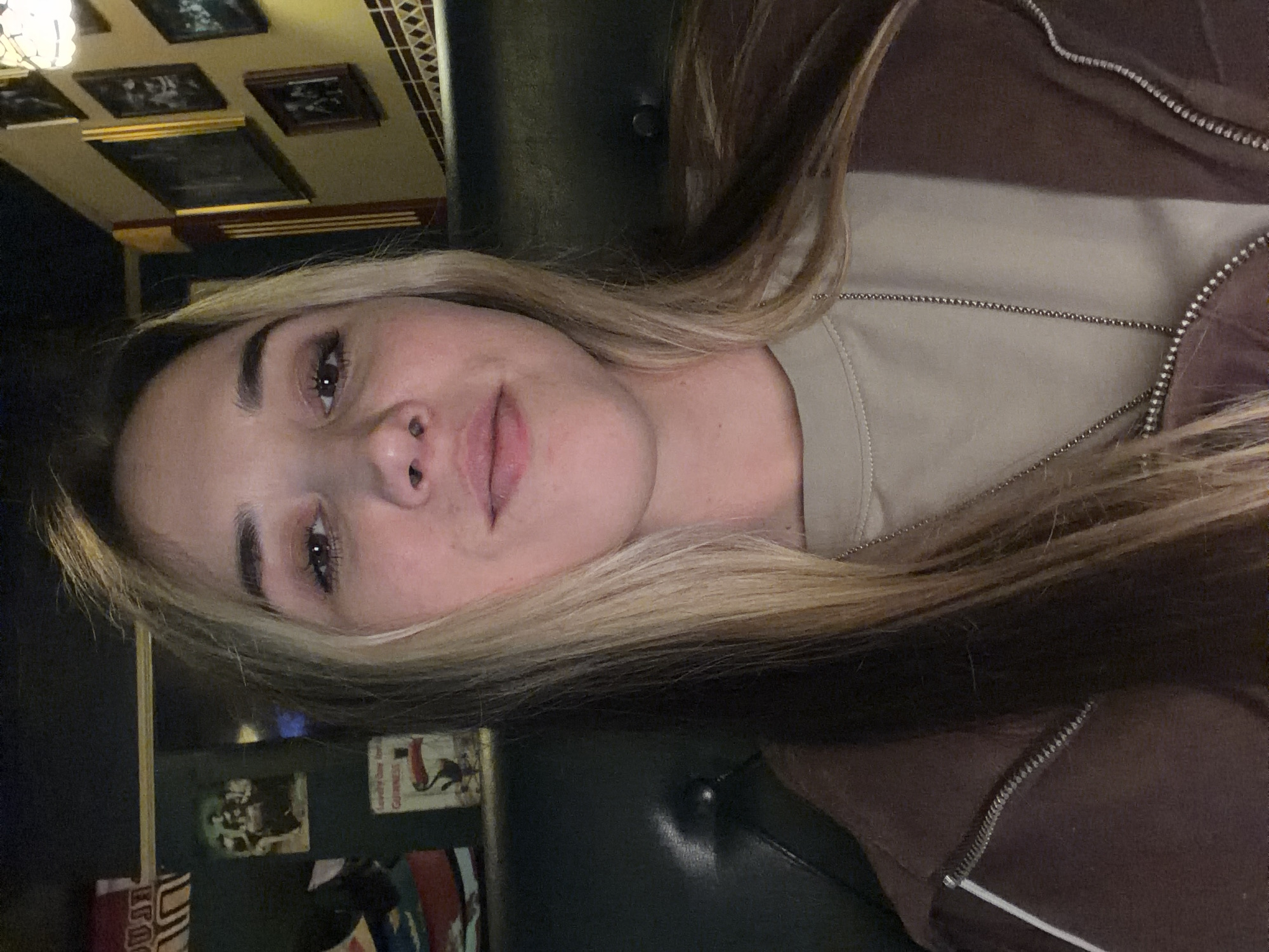 A woman with blonde hair is seated in what appears to be a restaurant or bar. She is wearing a brown and beige jacket, possibly a bomber-style jacket. Her face is clearly visible, showing light skin, brown eyes, and a nose piercing. In the background, there are framed pictures on the wall and what seems to be a leather chair or booth she is sitting on. There is no indication that the location is in Rostov-on-Don, Russia.