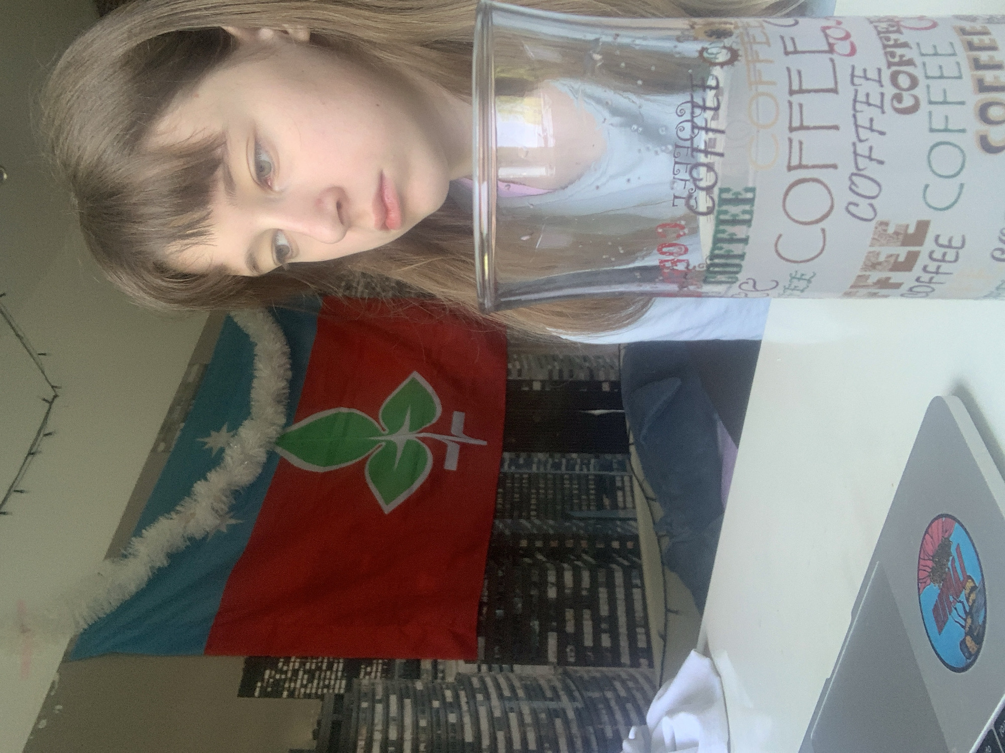 A woman is visible in the image, partially obscured by a glass printed with the word "COFFEE". Behind her, there is a flag with a blue and red background and a green plant symbol on it. Below the flag is a cityscape image. A laptop with a sticker is on a white table. The location cannot be confirmed.