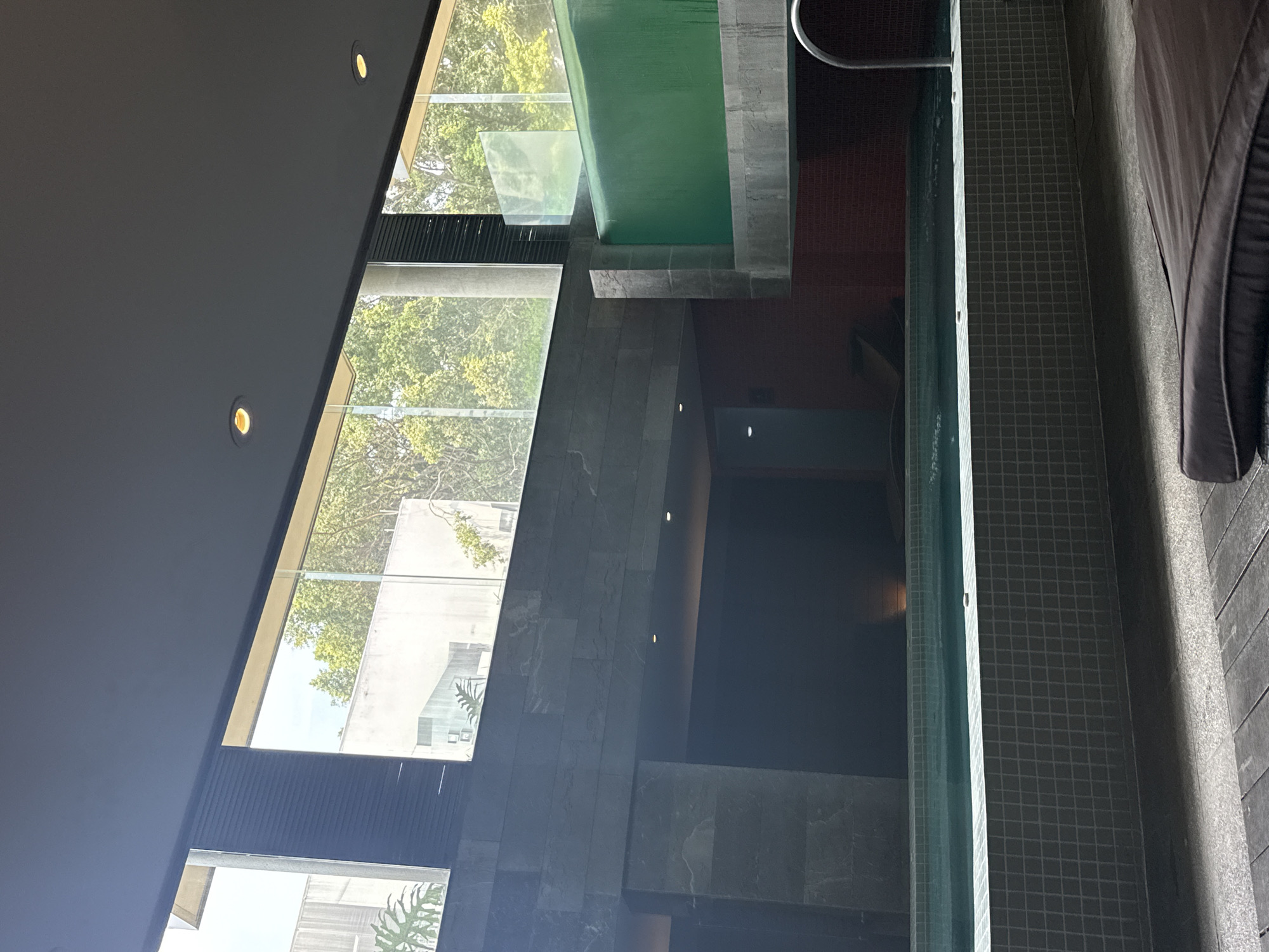This indoor shot, taken in Mexico City, Mexico, presents a partially obscured perspective of a multi-level indoor space. The focal point is a high-rise window that offers a view of exterior trees and a light-colored building. To the right, there is a tiled pool, next to a seating area, and a second higher-level pool is visible from the window. The lighting is predominantly natural daylight, supplemented by artificial ceiling lights. The overall impression is of a modern, architectural space with a mix of indoor and outdoor elements.