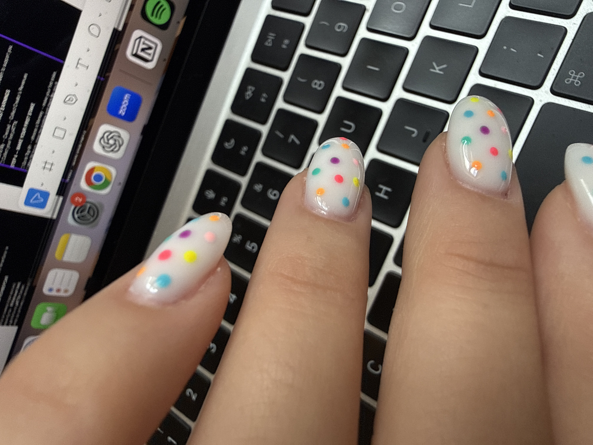 The image shows a close-up of a hand with manicured nails featuring a white base with colorful polka dots. The hand is positioned over a laptop keyboard, with app icons visible on the side of the screen. The keyboard and laptop suggest an indoor environment, possibly an office or home workspace.