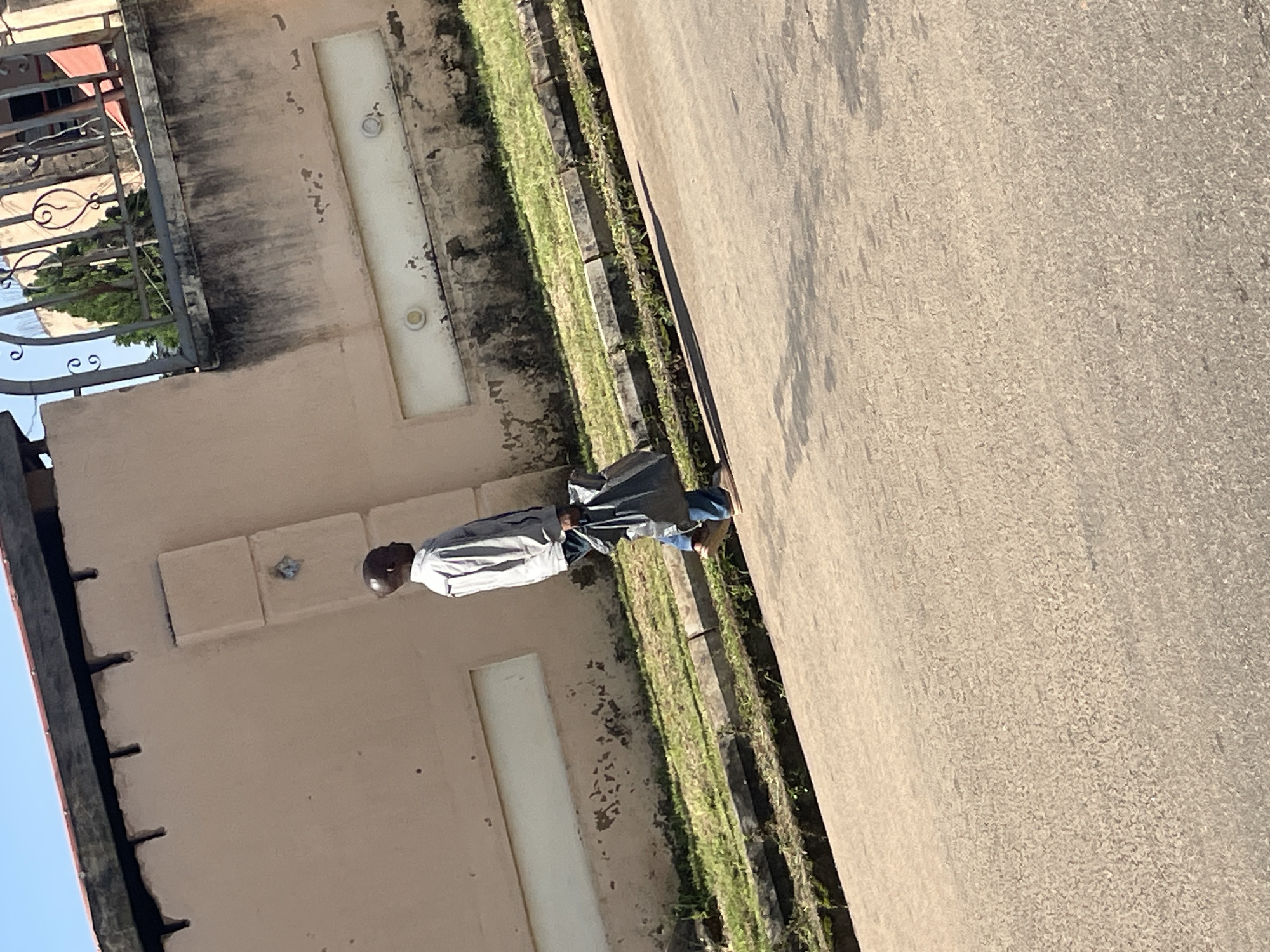 In Jos, Nigeria, a man is captured walking from left to right on a paved road. He is dressed in a light-colored collared shirt and blue jeans, carrying a black plastic bag in his right hand. Behind him, a grassy area is bordered by a concrete curb. Further back, a light-colored building with a textured facade stands, featuring a decorative horizontal panel with two circular elements. The building's roofline is visible, with visible wooden beams. A decorative metal fence is present in the background, with greenery and a structure visible beyond it. The sky above is clear and blue.