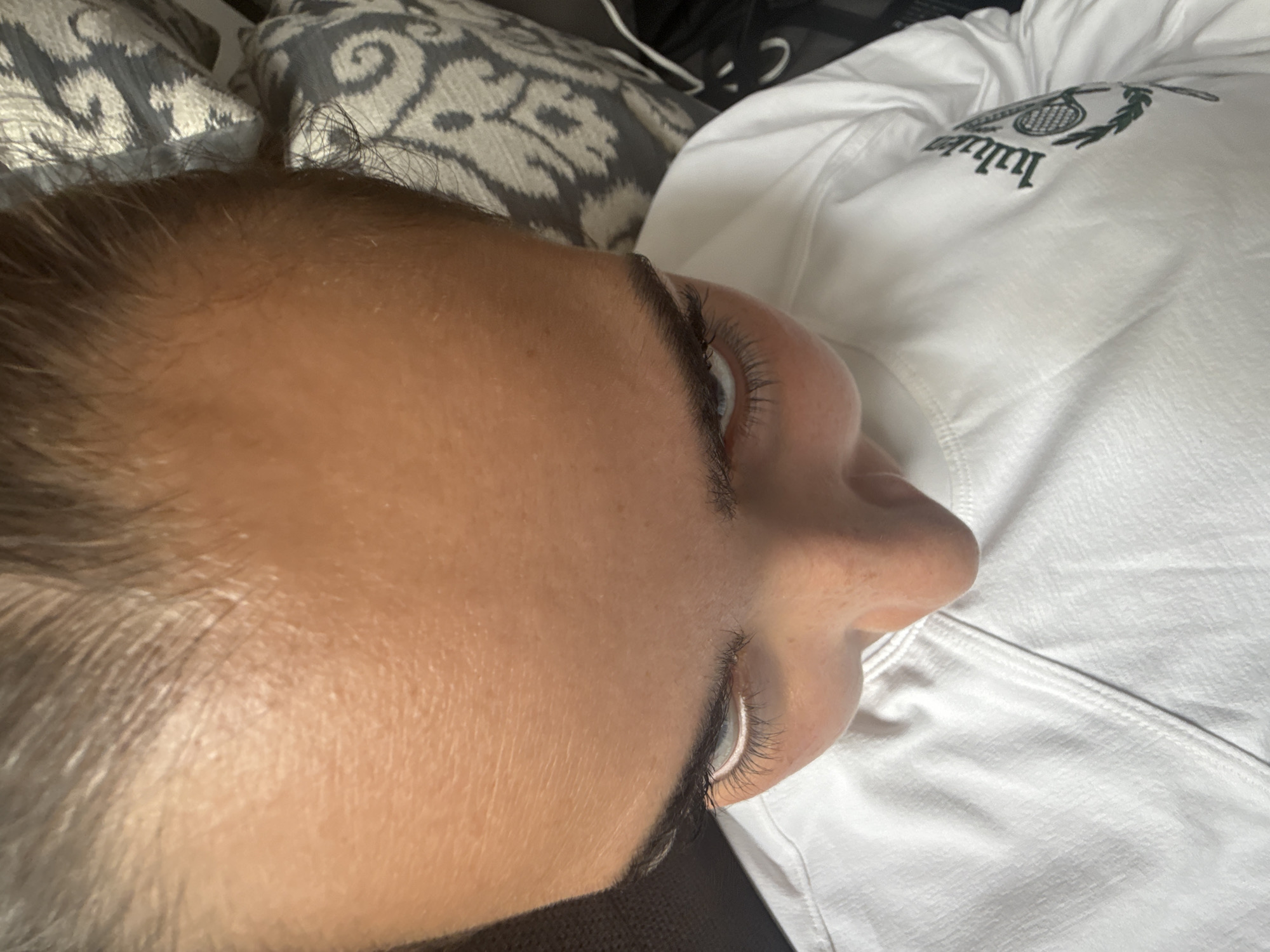A close-up, indoor, slightly angled shot depicts a person's face and upper body. The person's face is prominent, showing the forehead, eye, nose, and a portion of the cheek and hair. The person appears to be lying down, with their face tilted upwards. A white hooded sweatshirt with a logo is visible in the upper right portion of the image. Behind the person's head is a pillow with a grey and white patterned cover. The location is not identifiable as San Bernardino Tlaxcalancingo, Mexico, from the visual information provided.
