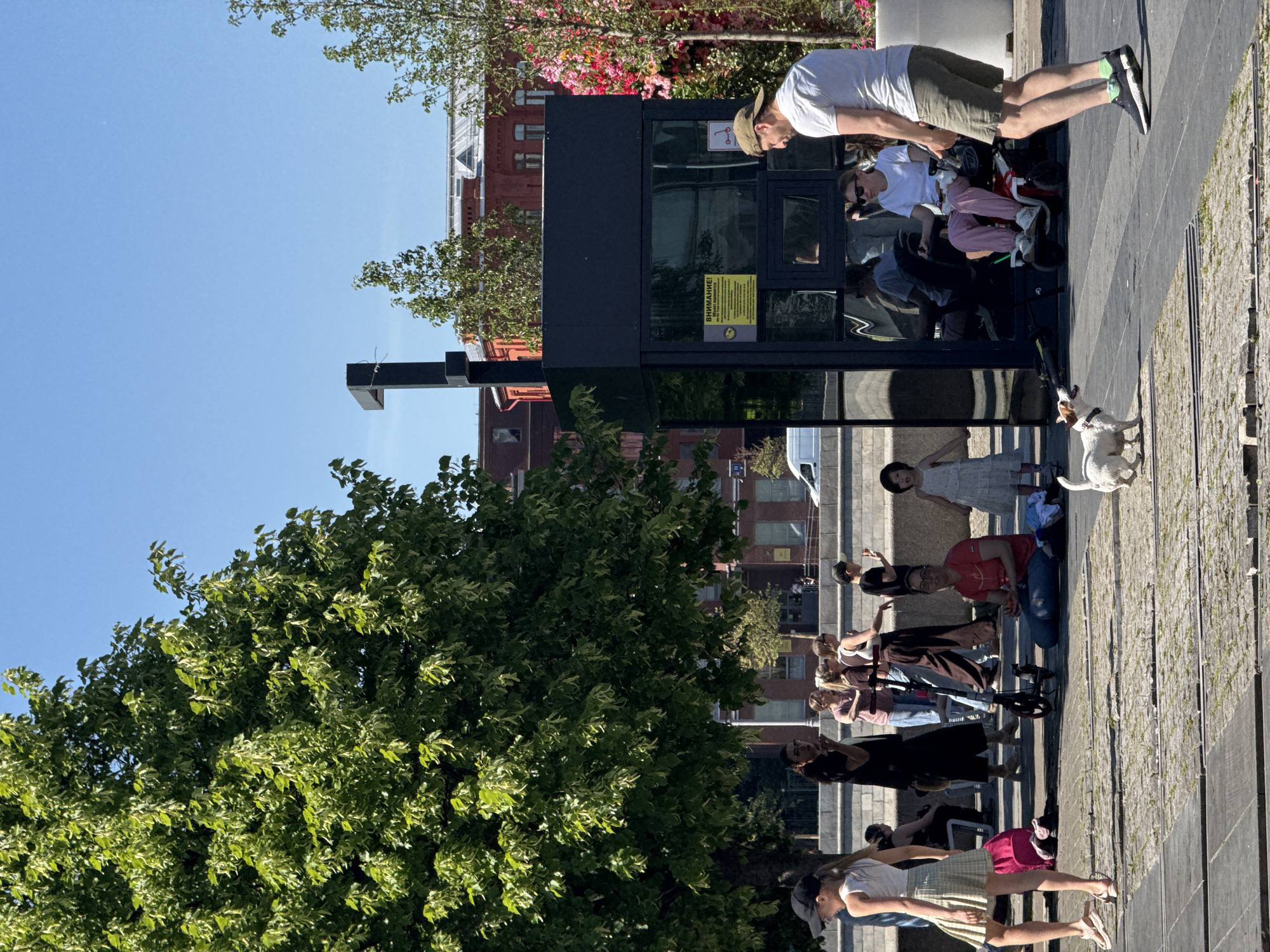 This detailed image captures a sunny outdoor scene along the Moskva River embankment in Moscow, Russia.
In the foreground, a young girl with light brown hair in a ponytail, wearing a black baseball cap, white t-shirt, and a light plaid pleated skirt, stands on the left, looking right. Her left foot is raised. To her right, a small white Jack Russell Terrier-type dog with brown patches and a black collar walks on the paved surface, looking up and slightly right, seemingly at a man on the right. This man, wearing a light-colored baseball cap, white t-shirt, and olive green shorts with black sneakers, walks towards the left, his gaze directed downwards, possibly at the dog.
In the mid-ground, a modern, black, glass-paneled kiosk or rental station stands prominently on the right. A yellow sign with Russian text "ВНИМАНИЕ!" (Attention!) and warning symbols is affixed to one of its glass panels, alongside a small kick scooter icon. An L-shaped black light fixture extends from the top of the kiosk. To the right of the kiosk, a woman in dark sunglasses, a white t-shirt, and pink pants is seated in a white, red, and black electric wheelchair, facing forward.
Further left in the mid-ground, a woman with glasses, wearing a red t-shirt and blue jeans, is seated on the ground, holding a bowl of strawberries. A small girl in a white dress stands beside her to the right. A black kick scooter lies on the ground nearby.
In the background, a large, lush green tree dominates the left side, providing shade. Below it, several people are visible: a woman with dark hair in a black outfit stands and looks right; others walk or stand casually, including one individual pushing a baby stroller. Beyond these figures, a low concrete wall suggests a riverbank. Across the river, or further back in the urban landscape, red brick buildings with white window frames and distinctive rooflines are visible under a clear, bright blue sky. A white van is also partially visible on the far left. The ground is paved with large, irregular stone blocks, with grass growing in the cracks. The scene depicts a lively public space on a bright day.