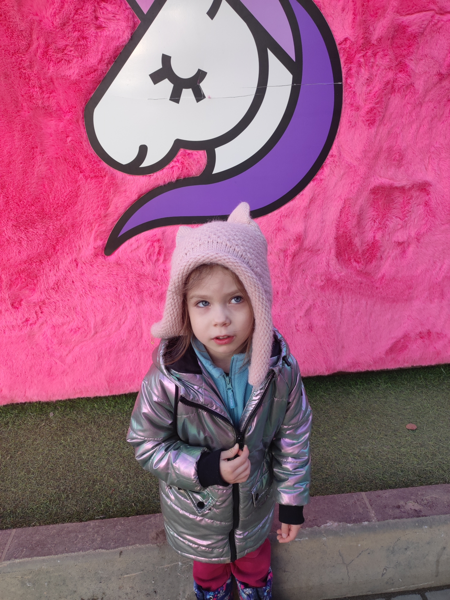 A young child stands in front of a large, pink, furry backdrop in Votkinsk, Russia. The backdrop features a cartoonish image of a white unicorn head with a purple mane. The child wears a pink knit hat with ear flaps, a silver metallic puffer jacket over a blue fleece, and patterned leggings. The child is looking upwards and to the left. The ground consists of artificial green grass and a concrete curb.