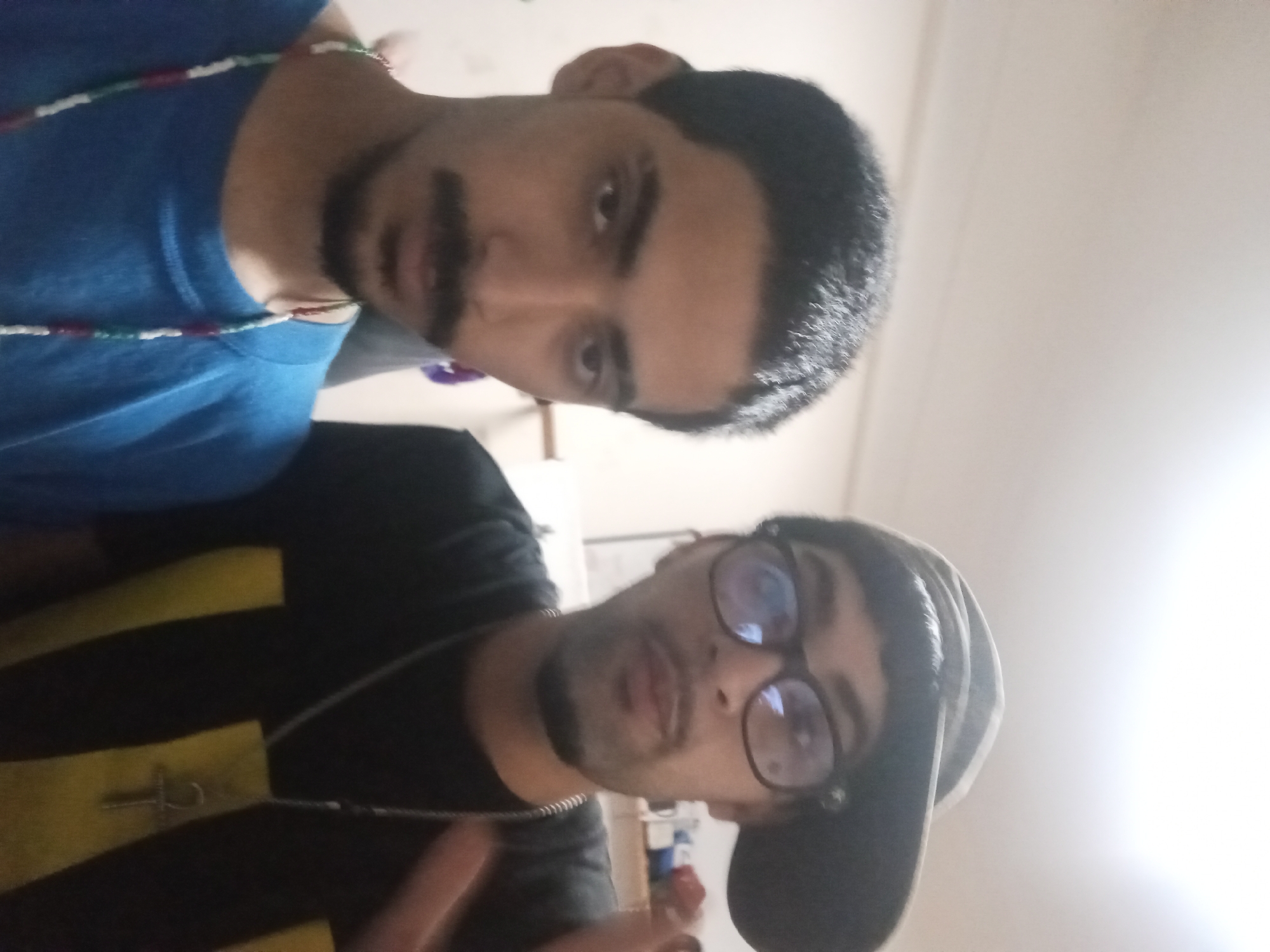 The image depicts two men in an indoor setting. One man is wearing a blue t-shirt and a beaded necklace. The other man is wearing a black t-shirt with yellow stripes, a necklace with an ankh pendant, and a black baseball cap. He also wears glasses. The location and city information are not discernible from the image.