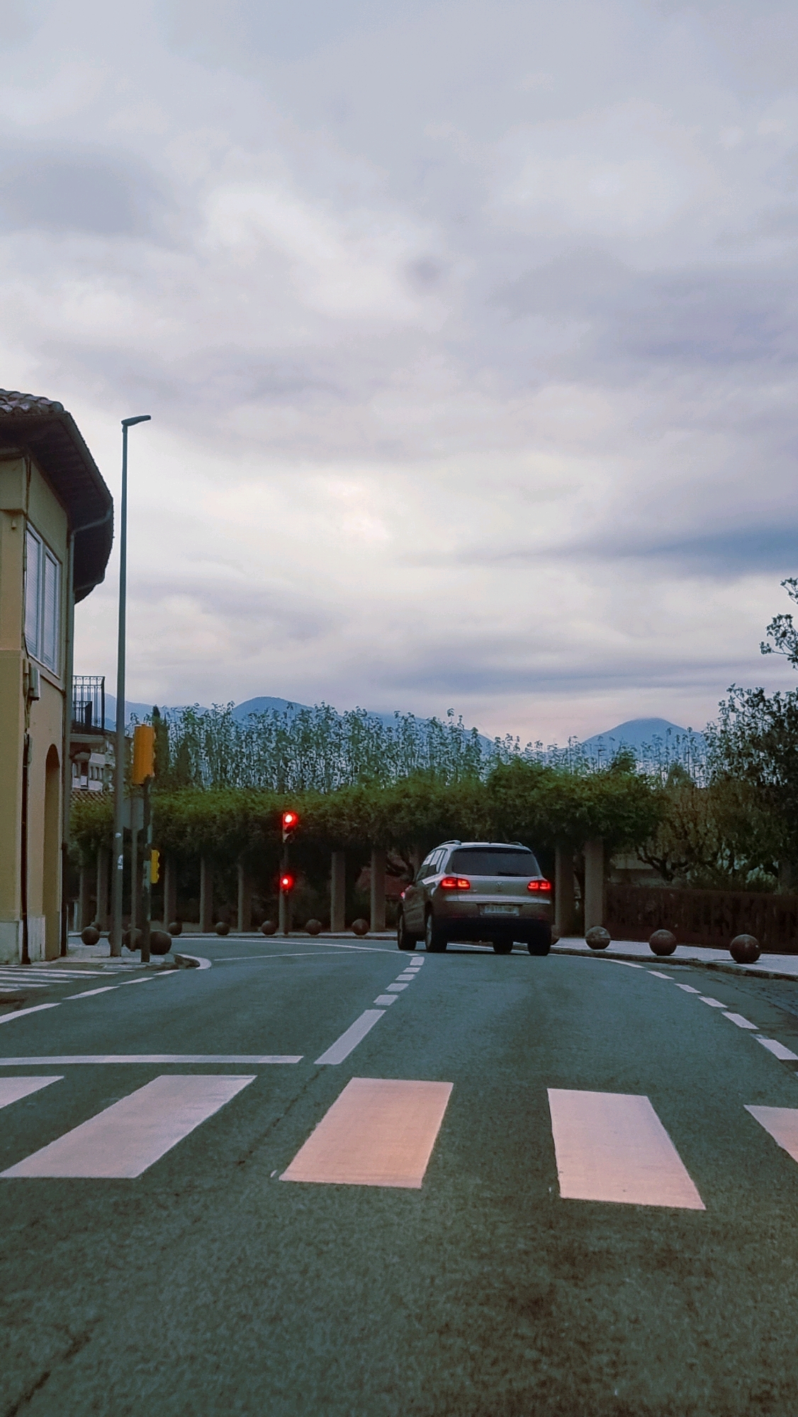 The image depicts a street scene in Sant Joan Les Fonts, Spain. A silver SUV is stopped at a traffic light, which is red. The car is facing away from the camera and is positioned on the right side of the road. In the foreground, a crosswalk is visible with white stripes painted on the asphalt. To the left of the road, a building with beige and brown architectural elements stands. A street lamp is also present on the left. In the background, a line of trees with dark green foliage is visible, and beyond that, mountains are seen under a cloudy sky. The overall lighting suggests it is either dawn or dusk.
