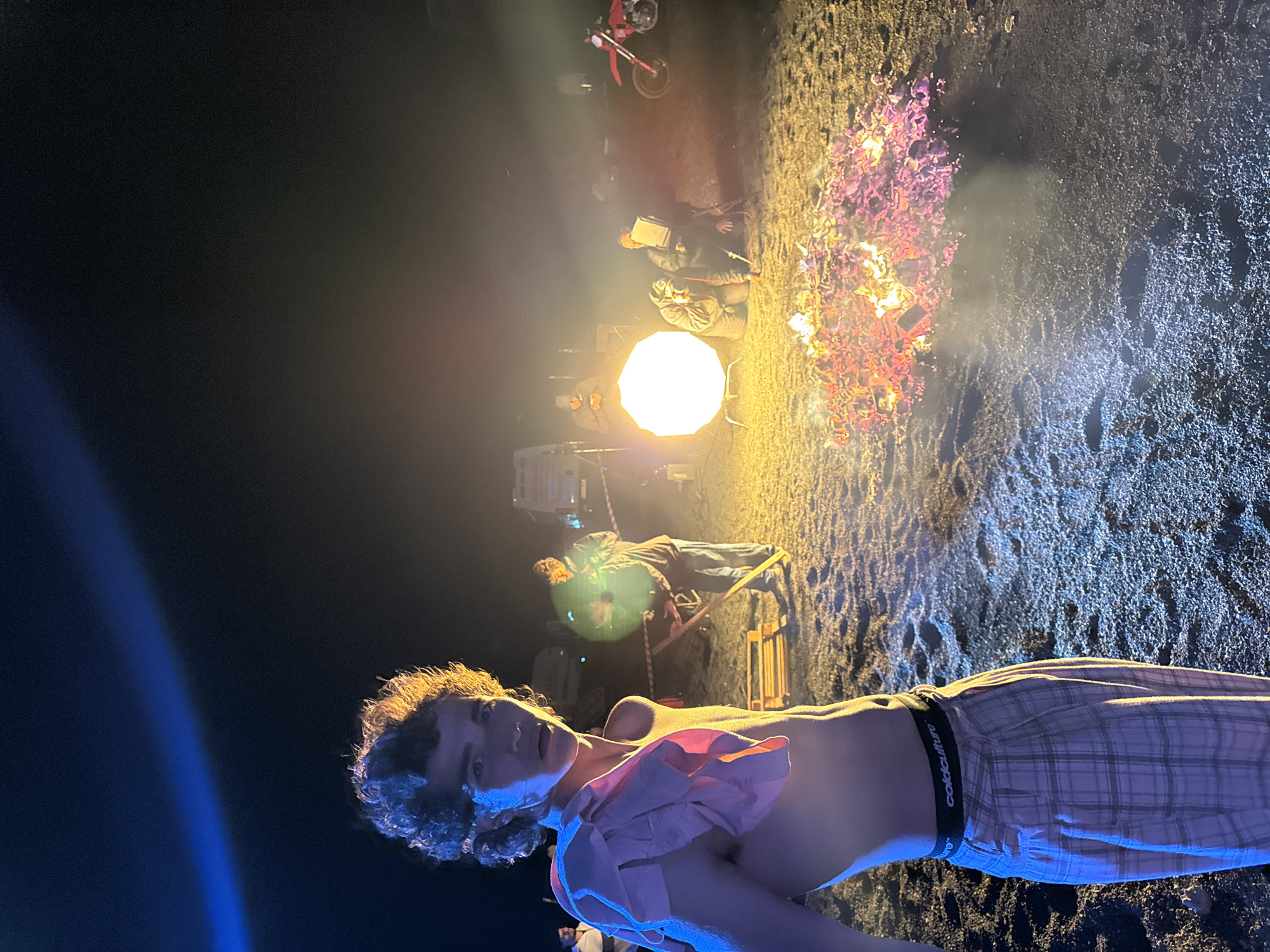 The image depicts a scene on a beach at night. A shirtless man in checkered pants stands in the foreground, illuminated by blue light. In the background, a bonfire burns, casting light on a group of people and equipment, including a large light source, chairs, and what appears to be a motorcycle. The ground is dark and grainy, resembling volcanic sand or ash. The image's context suggests it was taken in the municipality of Teguise, Spain, known for its volcanic landscapes and coastal areas. The overall atmosphere hints at a possible film set or organized event taking place on the beach.