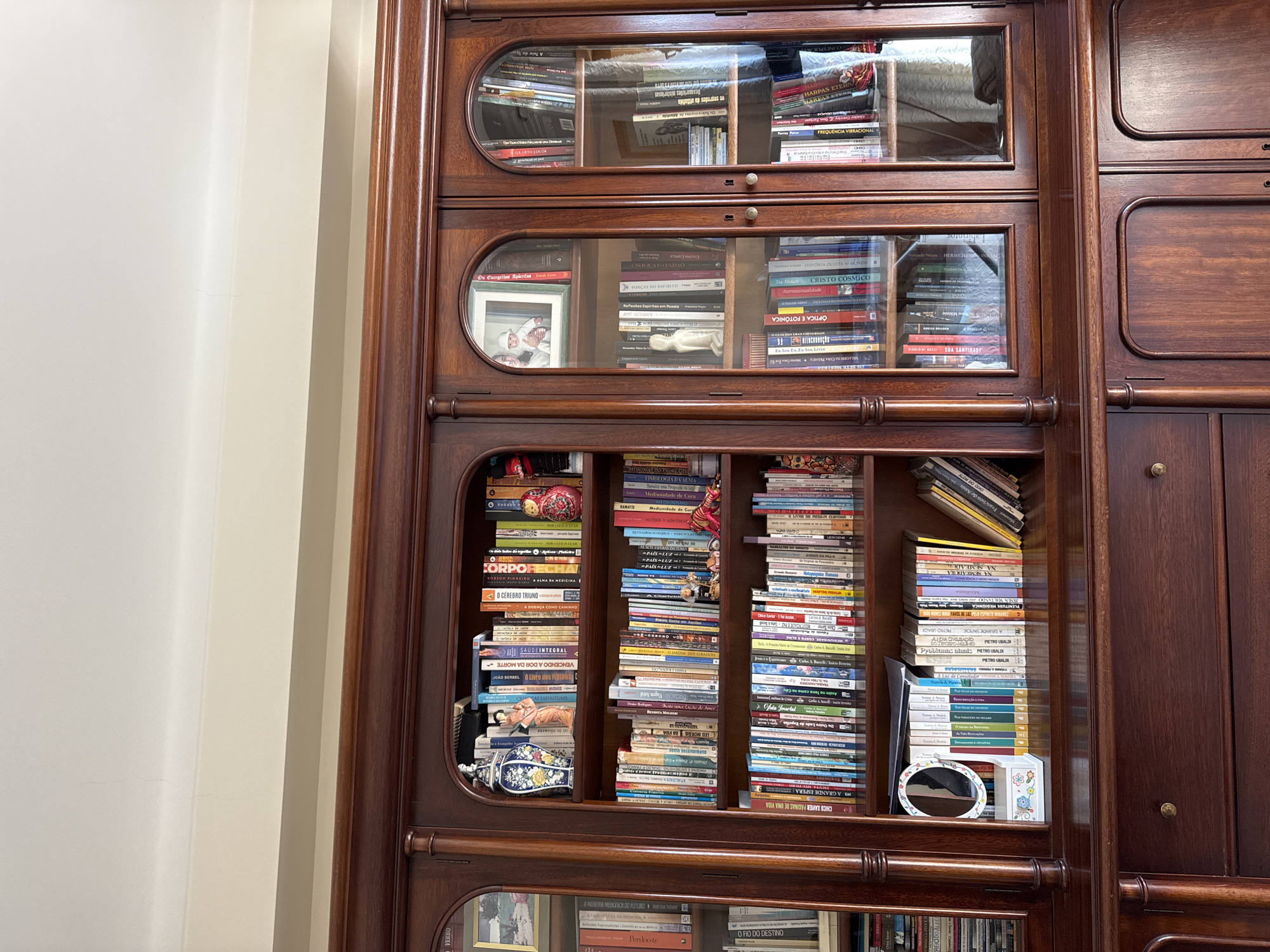 The image displays a section of a large, dark-stained wooden bookshelf and display cabinet located indoors in Mirandela, Portugal. The central portion of the cabinet features open shelving abundantly filled with books and decorative items, including a blue and white patterned vase, a figurine in traditional Japanese attire, and a red matryoshka doll. A small white framed mirror on a stand is also present on a lower open shelf. To the right, two sections with glass doors showcase more books, a framed photograph of an adult holding a baby, and a white figurine. The bottom of the cabinet consists of dark wooden drawers and solid cabinet doors. Above the cabinet, a light-colored wall transitions to a white ceiling, exhibiting a faint hairline crack. No individuals are depicted, and no actions or interactions are occurring.