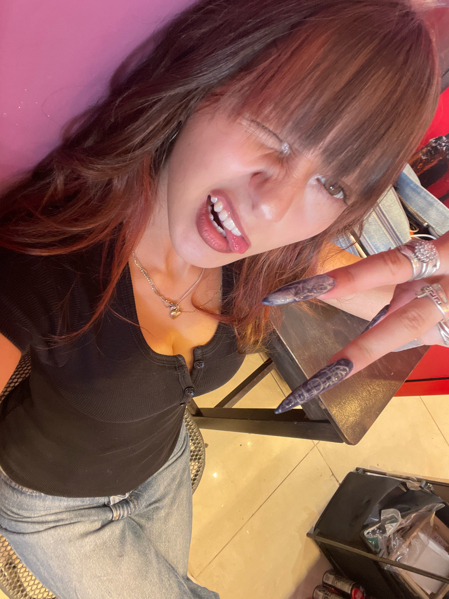 The image shows a person with long, reddish-brown hair, bangs, and an open mouth with their tongue sticking out. They are wearing a black, ribbed, button-up top and light-wash denim jeans. The person has long, pointed fingernails with a dark, possibly marbled, design and is wearing several silver rings. They are looking towards the camera, with their left eye squinted. In the background, a pink surface is visible on the left, and a table with a metallic surface is to the right. The floor appears to be tiled. A black cart with various items is in the lower right corner of the image. The scene appears to be indoors, possibly a personal space or a commercial establishment. The location is Raanana, Israel.