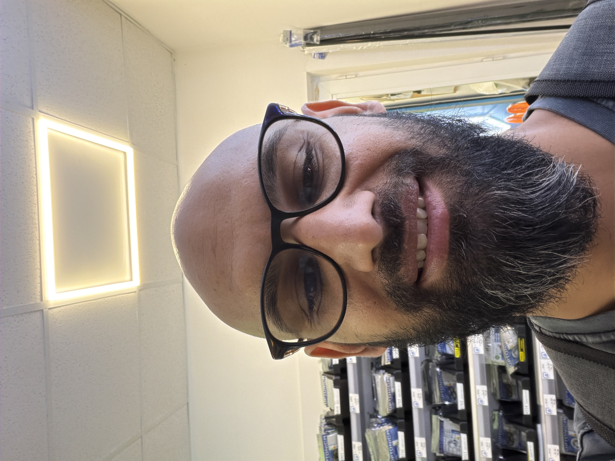 A close-up, selfie-style, medium-shot photograph captures a bald, middle-aged man with a full beard and black-rimmed glasses. He is looking directly at the camera with a slight smile, revealing his teeth. The man is wearing a grey t-shirt and a grey backpack.
Behind him, shelves are visible, stocked with numerous small, packaged items, some of which appear to be labeled with "OPTONICA." The background suggests an indoor setting, possibly a retail store or a pharmacy. The ceiling features a square, illuminated panel, and a portion of a door frame is visible to the right. The lighting is bright, casting minimal shadows.
This image was taken in Plovdiv, Bulgaria. The specific location within Plovdiv is unknown, but the presence of the stocked shelves and the overall environment suggests a commercial establishment.