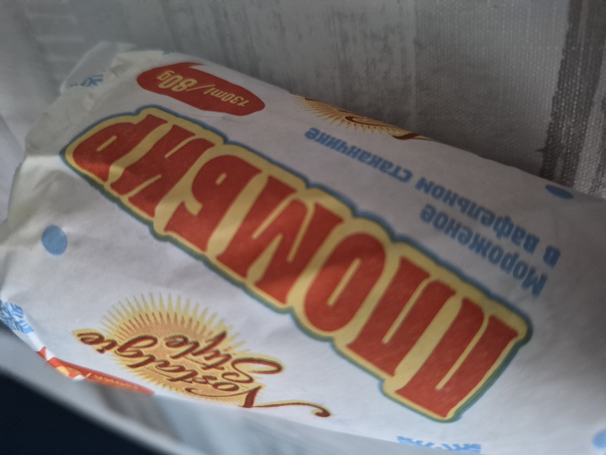 A close-up shot shows the packaging of an ice cream product. The packaging is predominantly white with bold, colorful lettering. The main text, in large red letters with yellow outlines, appears to spell "ПЛOMБИP" (Plombir), a type of ice cream. To the left, smaller text reads "Nostalgie Style" with a sunburst graphic. To the right, a red oval contains the text "130ml / 80g". Below the large text, in smaller blue letters, are the words "Мороженое в вафельном стаканчике" (Ice cream in a waffle cup). The product packaging is wrapped around an ice cream, obscuring most of its form. The background is blurred and appears to be a textured surface, possibly a tablecloth or fabric. The lighting is somewhat diffused, casting soft shadows. There are no people or specific city landmarks visible.