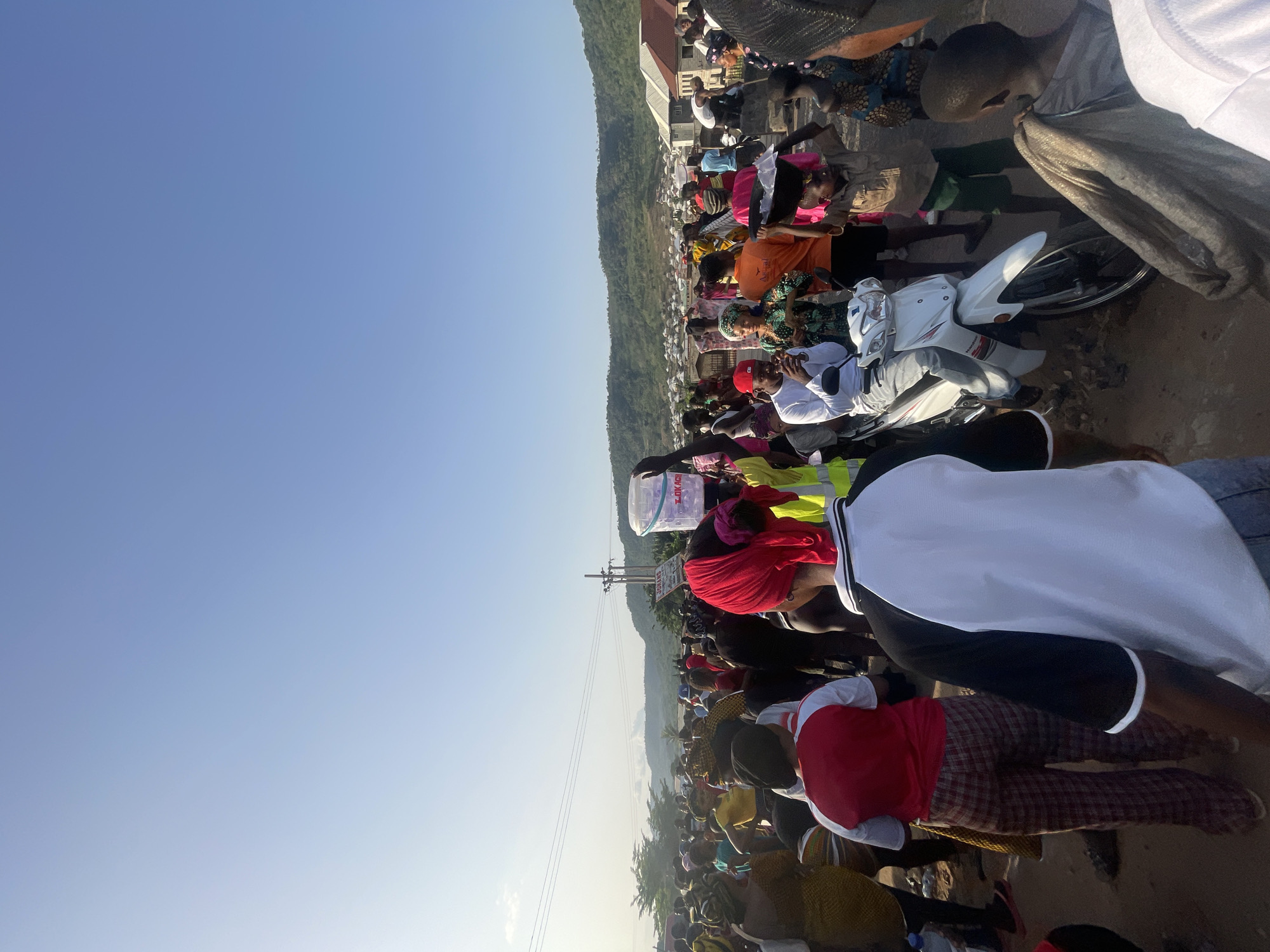 A horizontal outdoor shot depicts a large crowd gathered in what appears to be Lokoja, Nigeria. In the foreground and middle ground, numerous people are seen wearing various clothing, including red, white, and brightly colored garments. Some individuals are captured mid-action, while others are stationary. A white motorcycle stands out amidst the crowd. In the backdrop, buildings and a green hilly landscape are visible under a clear blue sky. Powerlines stretch across the scene.
