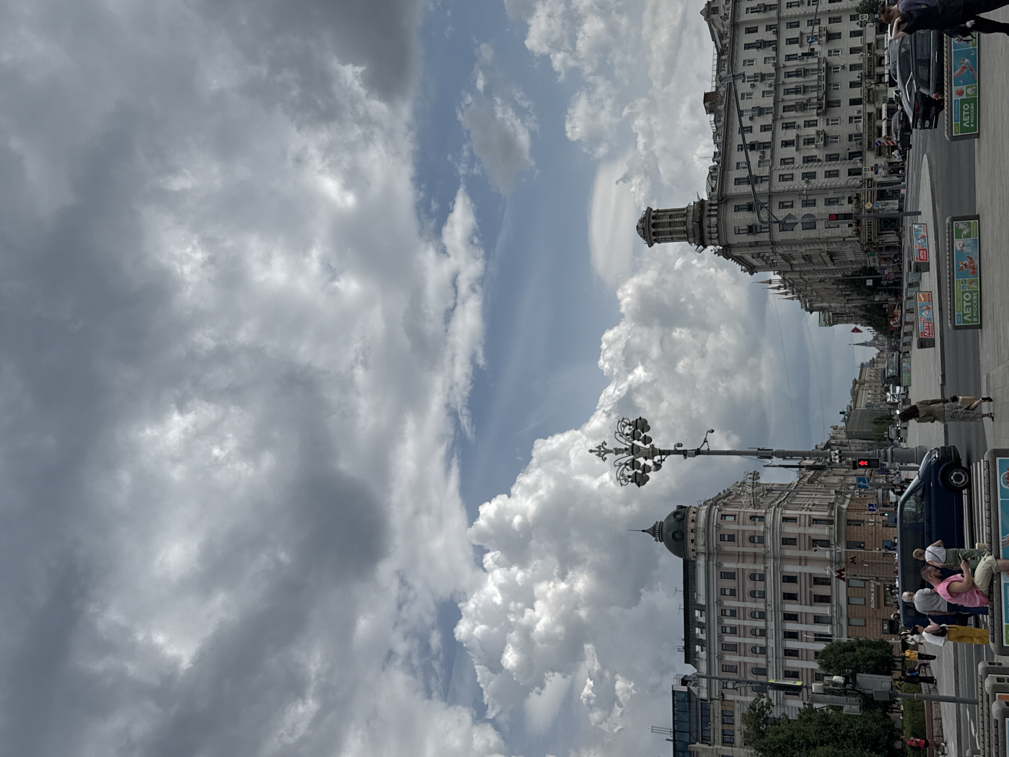 This is a full-frame, eye-level, outdoor shot of a city street in Moscow, Russia, under a dramatic sky filled with large, fluffy white clouds interspersed with dark, ominous storm clouds. The scene captures a wide, paved intersection with pedestrian crosswalks and traffic.
On the left, a multi-story building with a beige facade, arched windows, and a dark green dome tops one of its sections. A street lamp with multiple globes stands prominently in front of this building. Further down the street, more buildings with ornate architecture line the road, some with spires. On the right, a large, light-colored building with a rounded tower at its corner dominates the view, featuring numerous windows and decorative elements.
Several vehicles, including a dark blue minivan and black cars, are visible on the road. A traffic light displays a red signal. Numerous pedestrians are scattered throughout the scene, some walking, some gathered in small groups. A man in a pink t-shirt is seated on a low concrete barrier in the foreground, looking at his phone. Several advertising barriers with colorful illustrations are placed along the sidewalk. The overall impression is a busy urban environment preparing for or just after a weather event.