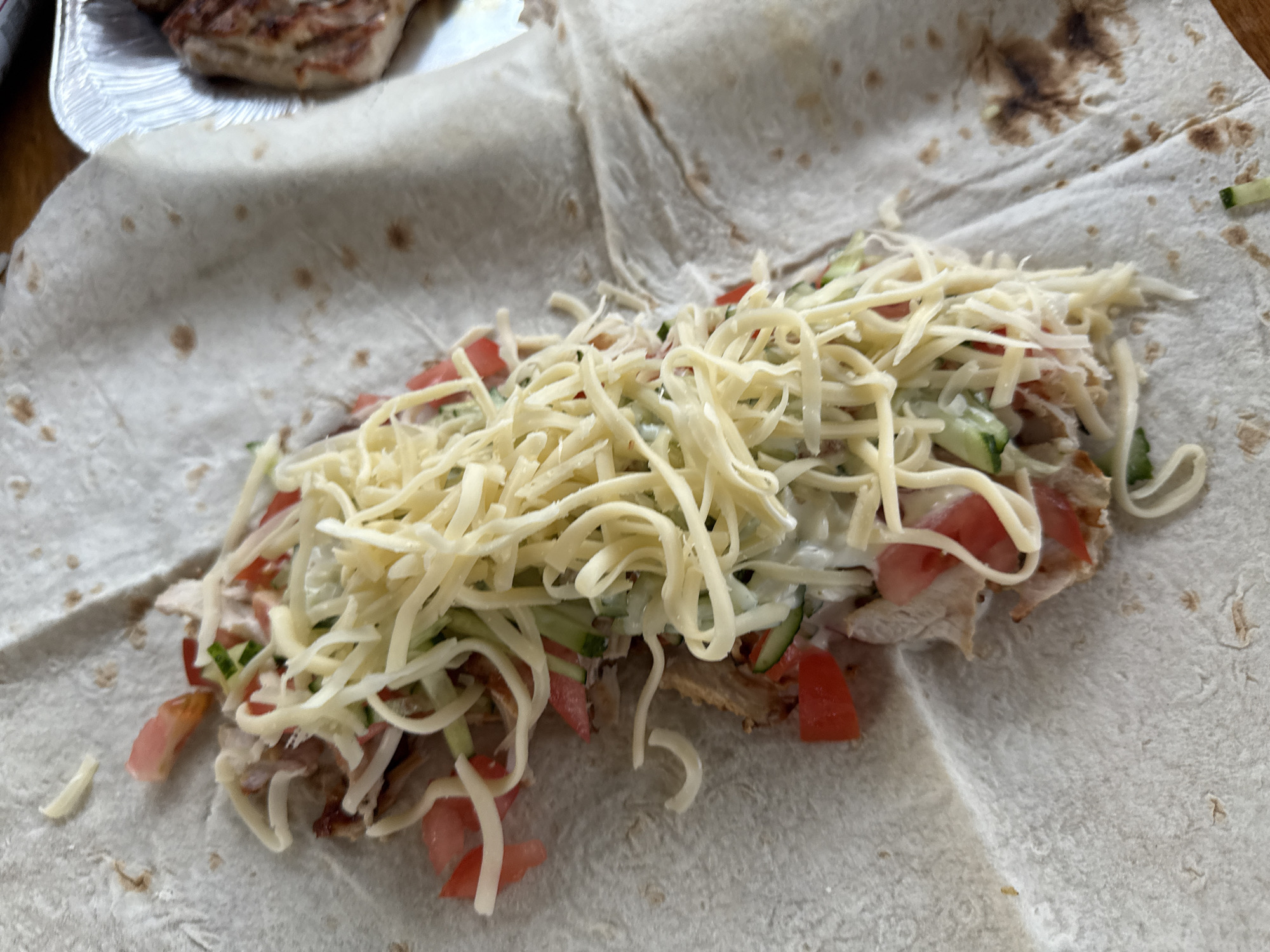 This media file shows a close-up, eye-level view of a half-assembled shawarma on a light-colored flatbread. The filling includes shredded cheese, chopped tomatoes, sliced cucumbers, and pieces of grilled chicken. The flatbread is spread open, with the filling concentrated in the center, suggesting the shawarma is in the process of being rolled. The background is blurred, but hints of a metallic serving tray with grilled meat are visible in the upper left corner, along with a wooden surface. There are no people, text, or weather cues visible. The lighting appears to be natural, likely from an indoor setting.