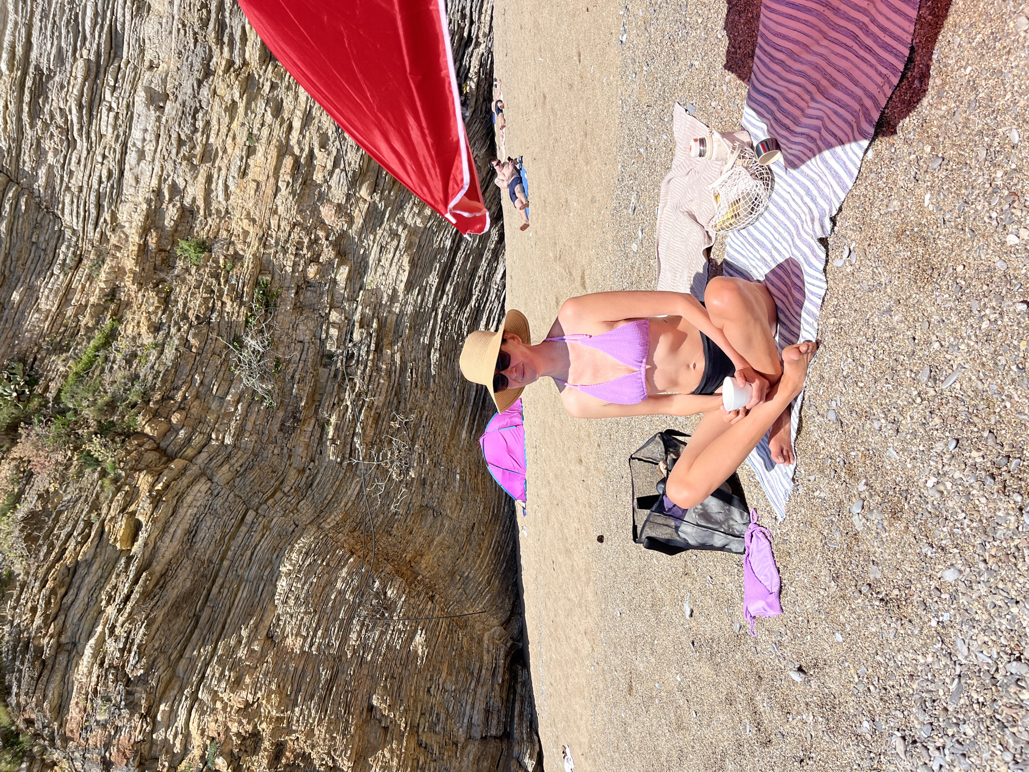 The scene is set on a beach, likely in Budva, Montenegro, given the rocky cliffs. A woman wearing a straw hat, sunglasses, a purple bikini top, and black bottoms is seated cross-legged on a striped towel. She is holding a white jar, possibly sunscreen. A bag sits near her, along with a purple bikini bottom. Behind her, a rocky cliff rises, partially shaded by a red sunshade or sail. In the background, other people are lying on the beach under the sun. A few beach accessories like a picnic basket and containers are on the towel. The beach is composed of sand and small pebbles.