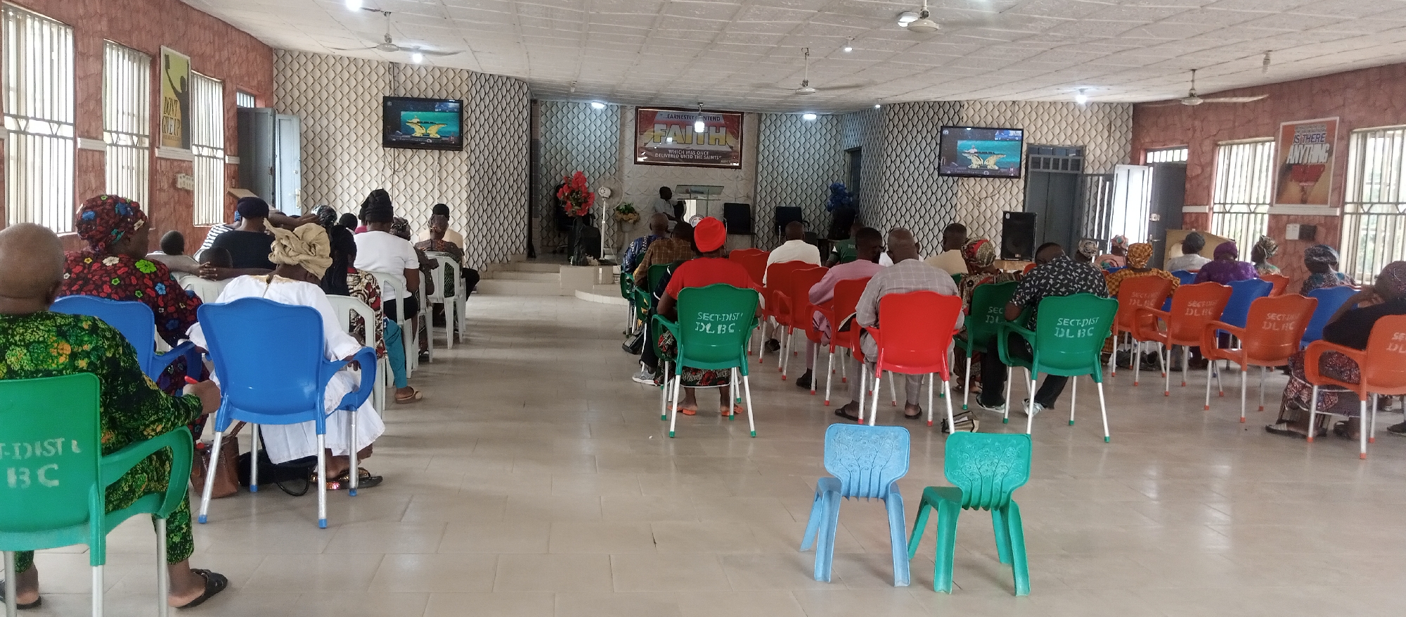 The image shows a gathering of people sitting in an indoor setting, likely a church or meeting hall, in Adankolo, Nigeria. Rows of colorful plastic chairs, mostly occupied, are arranged facing a slightly elevated stage or front area. A floral arrangement is visible on the stage. Two TVs mounted on the walls display images. Wall posters with text and images are also visible. The room is lit by ceiling lights and natural light entering through windows. The individuals are mostly adults, and some are wearing traditional attire.