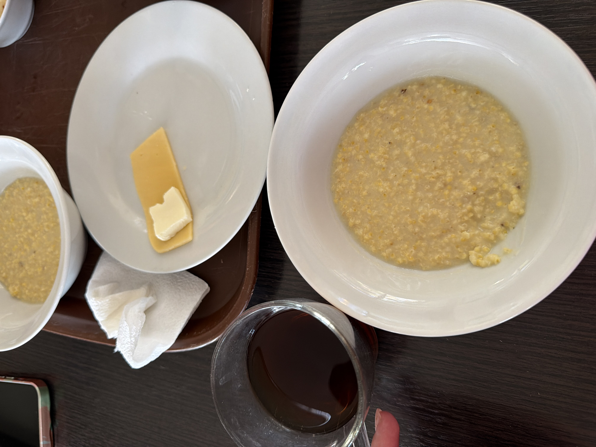 The scene depicts a breakfast setting, likely in Chaykovsky, Russia, based on contextual assumptions. A dark-stained table serves as the backdrop for a brown tray. The tray holds a white bowl of porridge, a white plate with a slice of cheese topped with a pat of butter, another bowl of porridge, and a wadded-up napkin. Next to the tray sits a glass of dark liquid, probably tea or coffee, and a portion of a cell phone is visible in the lower-left corner. The composition indicates a meal, presumably breakfast, is ready to be consumed. There are no people visible in the frame, but one finger is grasping the glass.