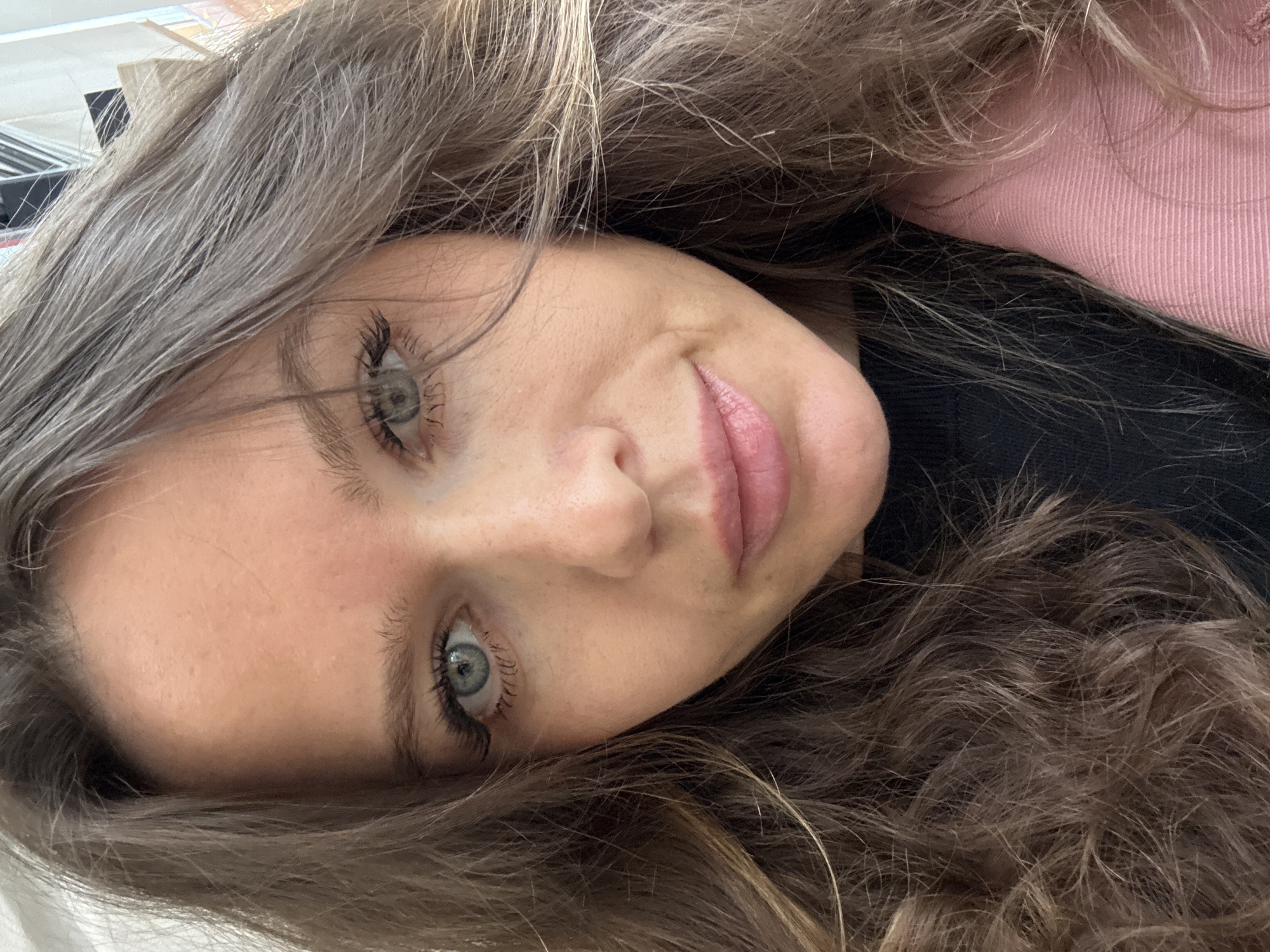 A close-up selfie shows a young woman with a calm and pleasant expression, her head tilted to the right, almost horizontal in the frame. Her long, wavy hair, a mix of light brown and auburn tones with subtle lighter streaks, cascades around her face and fills much of the image. She has striking light blue or grey eyes with prominent dark eyelashes, looking directly towards the camera. Her fair skin shows a few minor blemishes. Her lips are a soft pink, slightly parted in a gentle smile.
In the background, out of focus, a segment of a pink garment with a ribbed texture is visible behind her hair on the right side, adjacent to a darker, possibly black, garment or surface. The overall lighting suggests an indoor setting, though no specific time of day or light source is evident. The image does not contain any visible text. Based on the provided context, the location is Moscow, Russia.