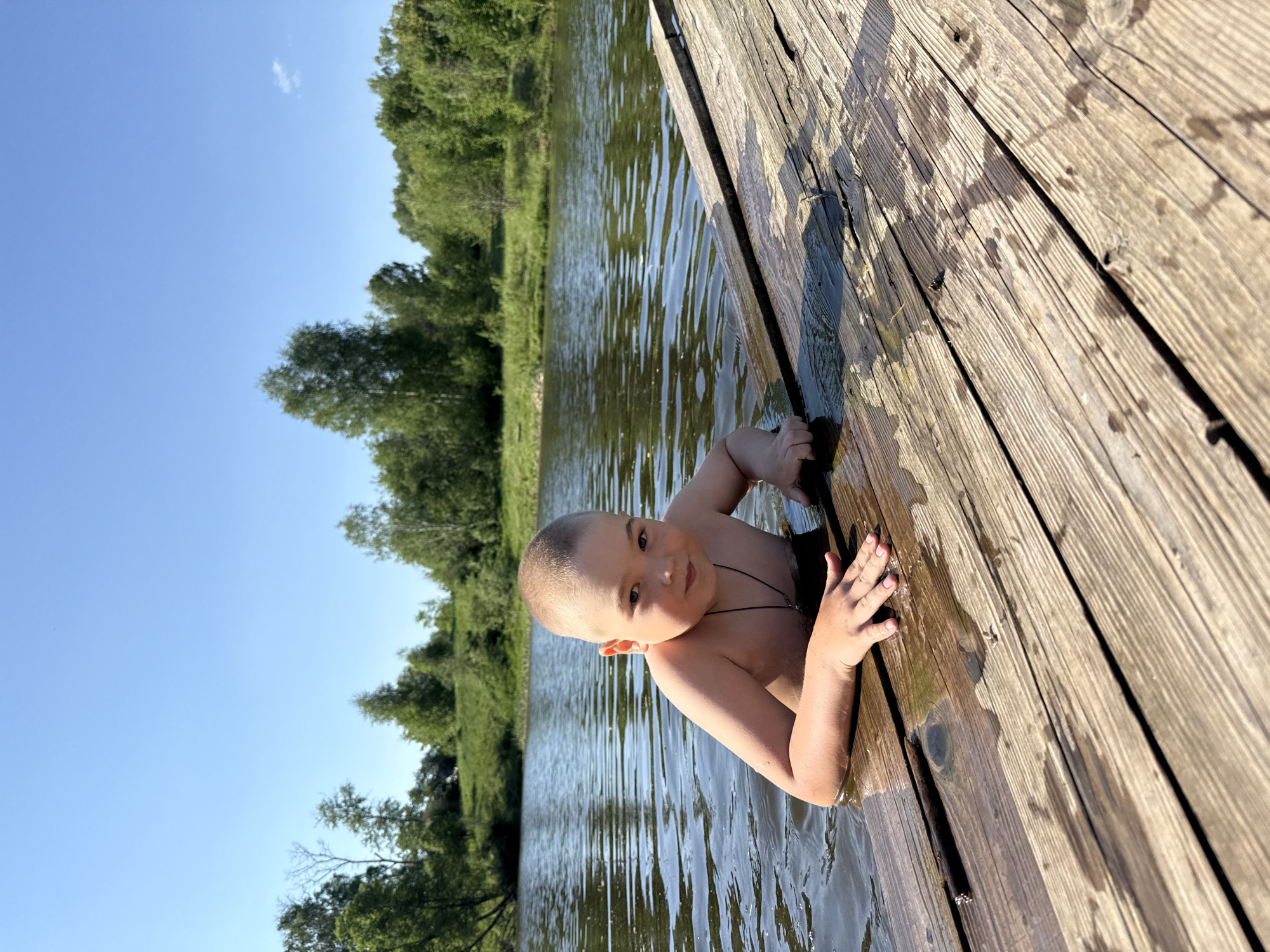 A young boy is emerging from a body of water, possibly a lake or river, onto a wooden dock. The dock's planks run diagonally from the lower right to the center of the image, with the boy positioned along its edge. He is gripping the dock's side with both hands, appearing to be climbing out. The water is rippled, reflecting the surrounding trees and sky. Trees line the opposite shore of the water body, and a clear blue sky is visible above. The location is Zhigalki, Russia.