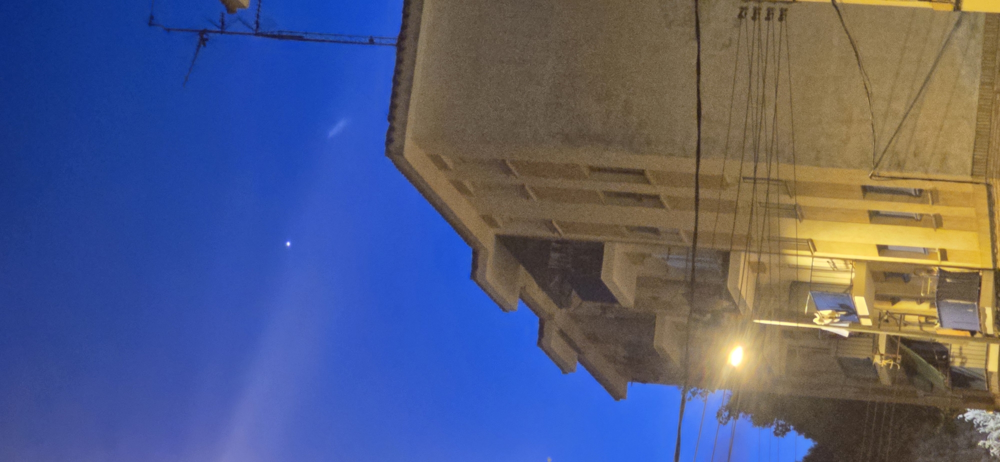 The image displays a tall, multi-story building with a beige facade, likely an apartment building, under a deep blue twilight sky. Several balconies are visible on the building's left side. A bright street lamp illuminates the lower portion of the building. Electrical wires are strung across the scene. In the upper part of the sky, a single bright star or planet is visible, and a faint streak of light, possibly a contrail, is also present. An antenna is mounted on the roof of the building on the right. There are no people clearly visible in the image. The environment appears to be an urban setting in Huesca, Spain, given the context provided.