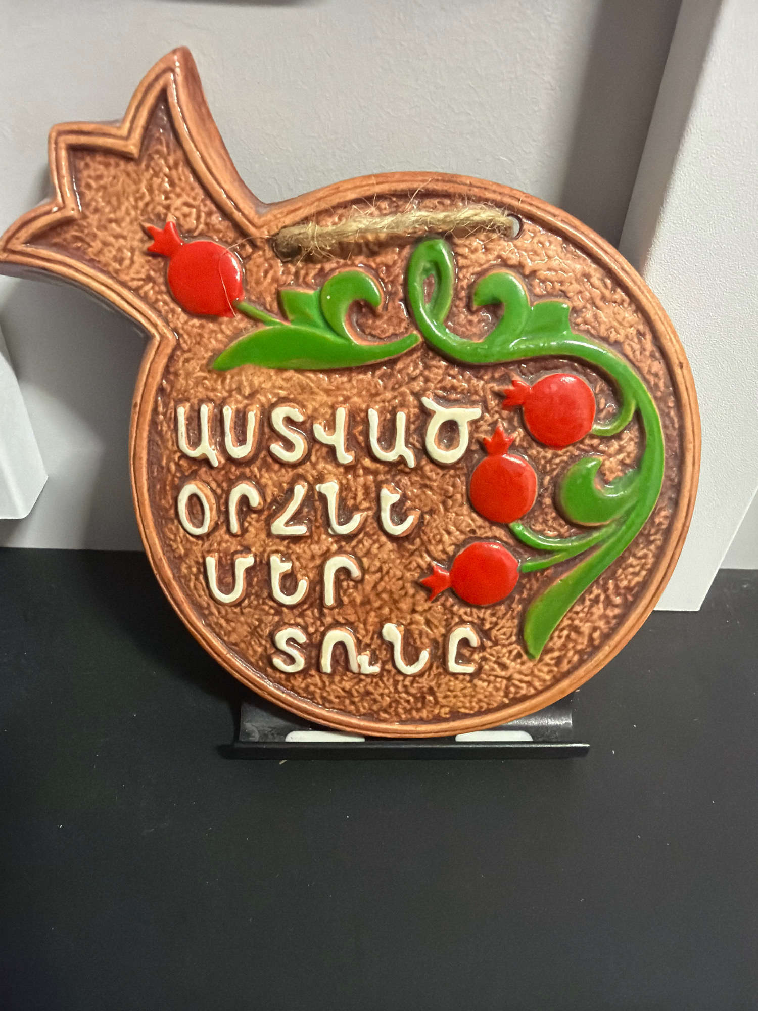 The image shows a ceramic pomegranate-shaped ornament with Armenian script. The ornament is brown with red pomegranates and green leaves. The Armenian script is white and appears to be raised from the surface. The ornament is hanging from a piece of twine. The background is a light gray wall. The ornament is positioned on a black surface with a black stand underneath. There is no discernible connection to the city of Vniissok, Russia.