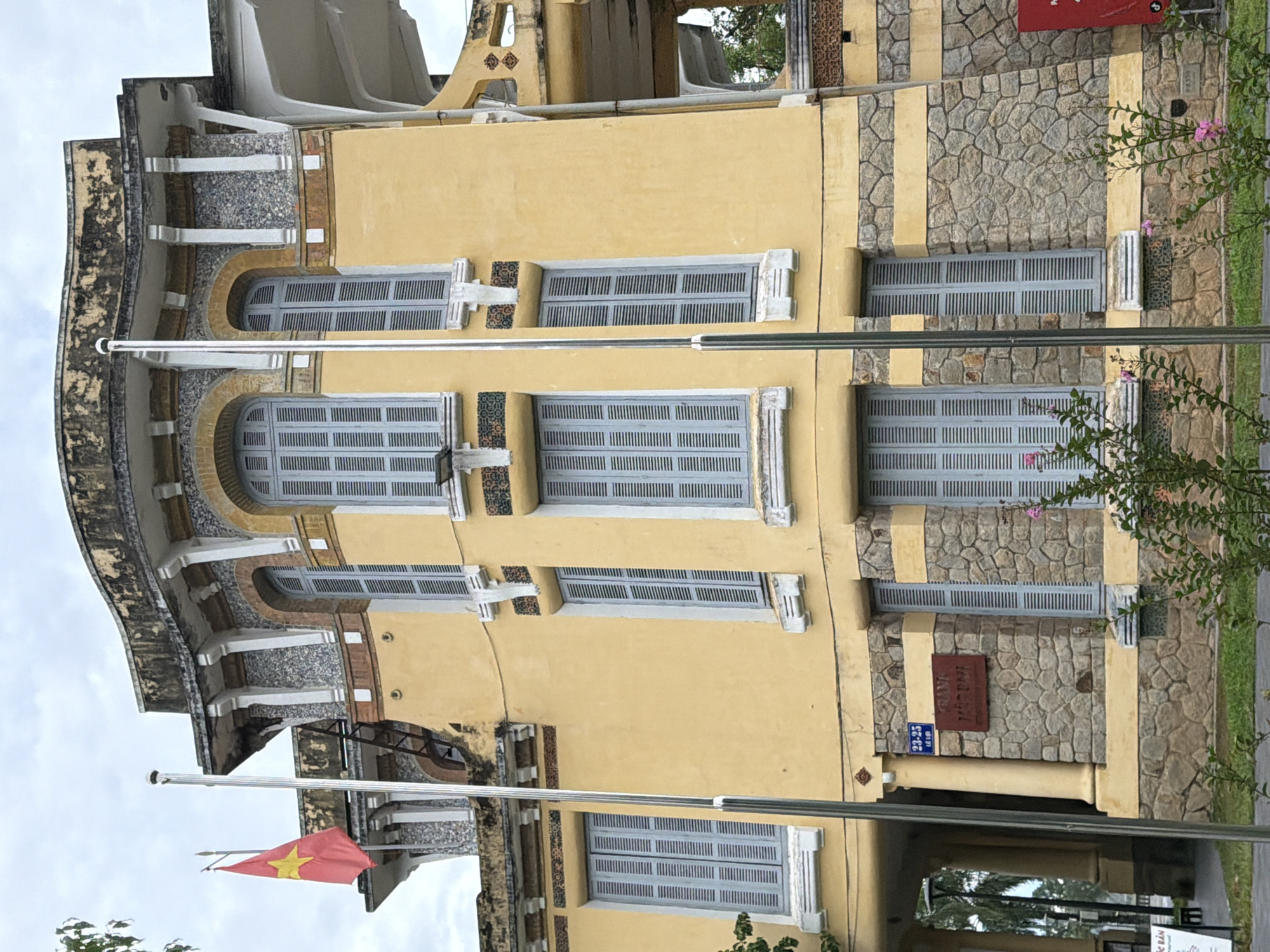 A yellow building with a stone base stands under a cloudy sky. A Vietnamese flag flies on a pole to the left of the building, which has multiple tall, shuttered windows and decorative stonework. A sign on the building reads "23-25 Le Loi" and another sign appears to say "TRANG MUSIC". The building is located in the city of Thuận Hóa Ward, Vietnam.