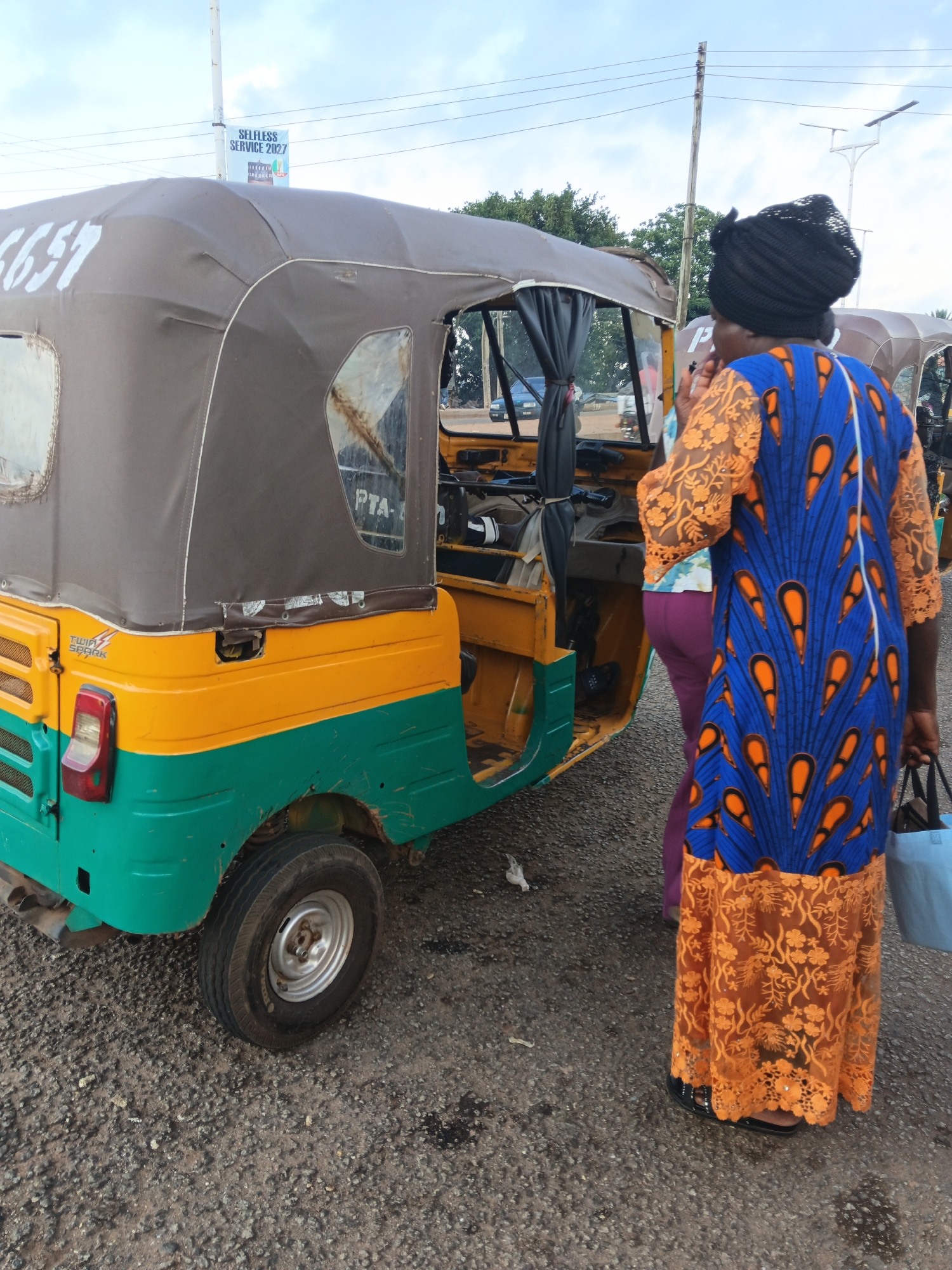 An outdoor, eye-level, medium shot captures a woman standing next to a three-wheeled auto-rickshaw in Bukuru, Nigeria. The auto-rickshaw is painted in yellow and green with a brown canvas top and white lettering on the side that reads "6657". The woman is wearing a dark headwrap and a vibrant blue and orange patterned dress with lace detailing at the hem. She is holding a light blue tote bag in her right hand. Another person, partially visible, stands in front of her, wearing purple leggings and a white and blue patterned top. The ground is covered in gravel. In the background, a utility pole with wires and a sign reading "SELFLESS SERVICE 2027" are visible. Trees and a cloudy sky complete the scene. The overall impression is of a typical street scene in a Nigerian city.