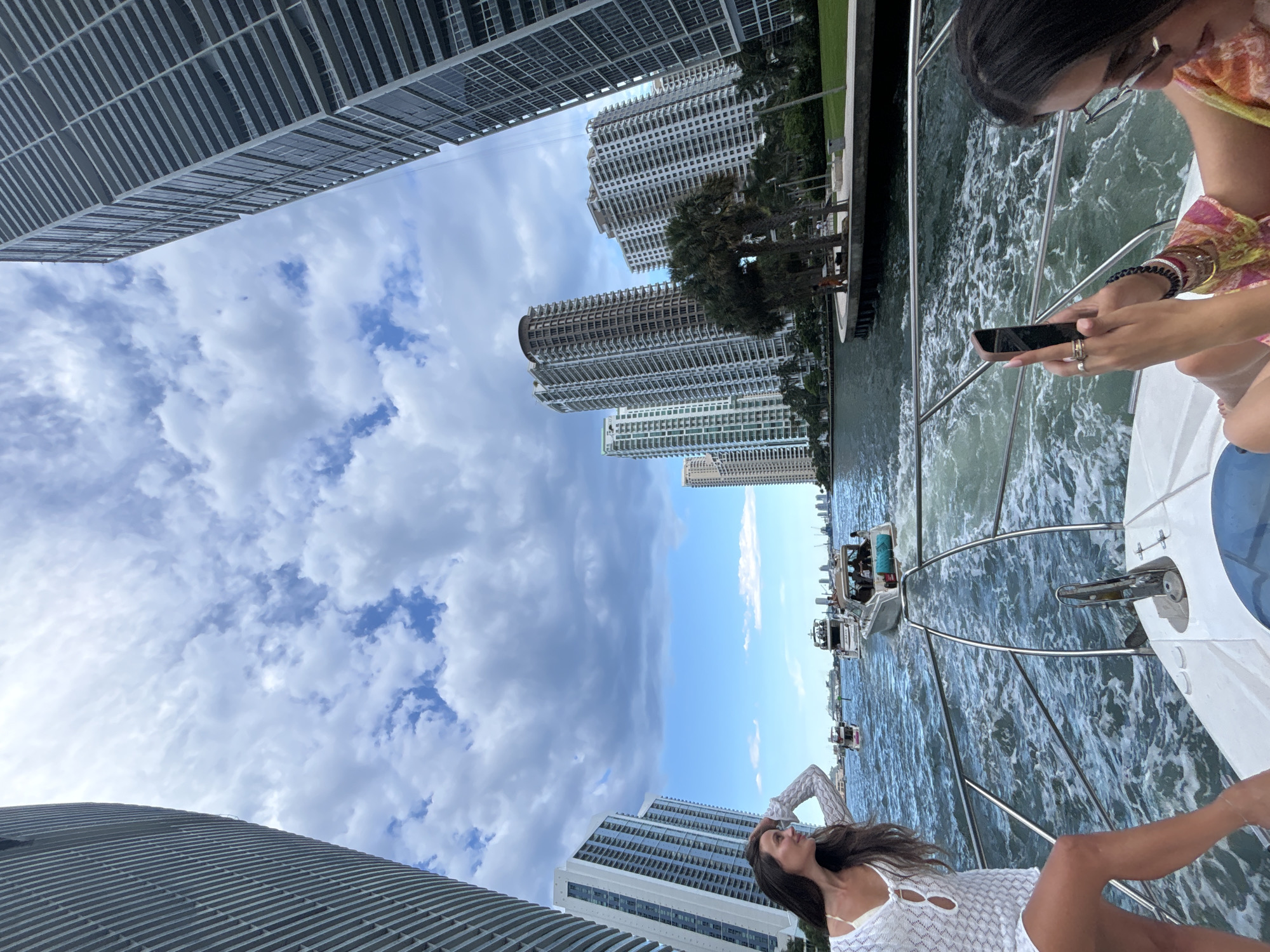 The image captures a sunny day in Miami, Florida, along the Miami River. A woman in a white cover-up is leaning on the railing of a yacht, looking towards the city skyline. She is wearing a white crochet cover-up over a bikini top and has long dark hair. Her left hand is raised to shield her eyes from the sun. In the foreground, the bow of a white yacht is visible, with churning water in its wake.
In the midground, several other boats, including speedboats and a larger yacht with passengers, are navigating the river. The cityscape features a dense collection of modern high-rise buildings with a distinct architectural style, some with balconies and glass facades. Palm trees are visible along the riverbank, suggesting a tropical environment.
In the right foreground, another woman, partially visible, is seated on the yacht, looking at her smartphone. She is wearing a colorful patterned top and glasses. The sky is partly cloudy with a mix of blue sky and white clouds. The overall scene depicts a luxurious boat tour or excursion in the urban setting of Miami.
