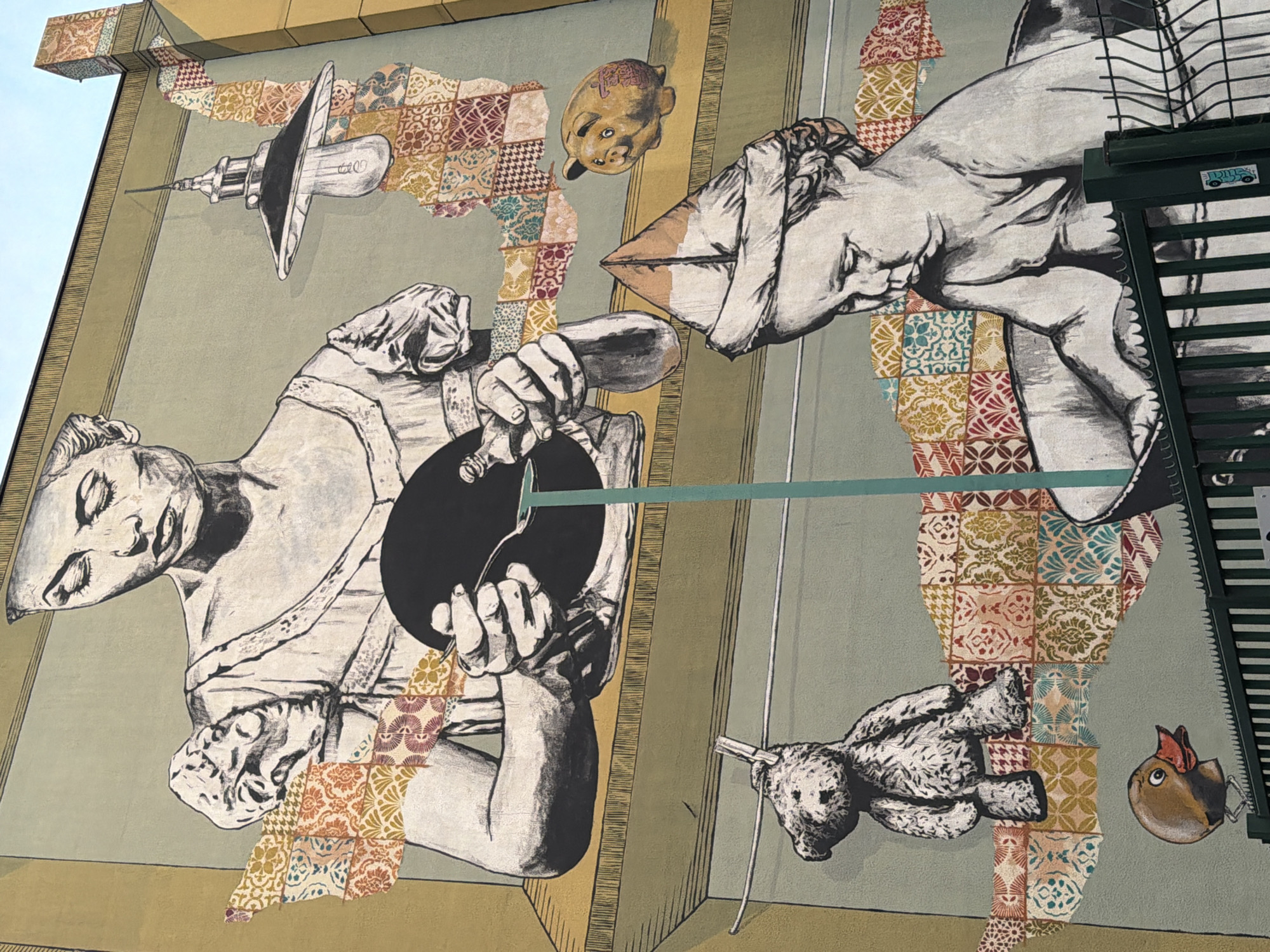 The image shows a mural on a building in Aubervilliers, France. The mural features a collage-like design with grayscale drawings of a figure pouring liquid into a dark circle, disembodied hands, a toy bear hanging by a string, and a lightbulb resembling a syringe. A yellow piggy bank is floating above the figures. The background of the mural is a patchwork of patterned tiles and vertical lines. The edge of a fire escape is visible on the right side of the image.