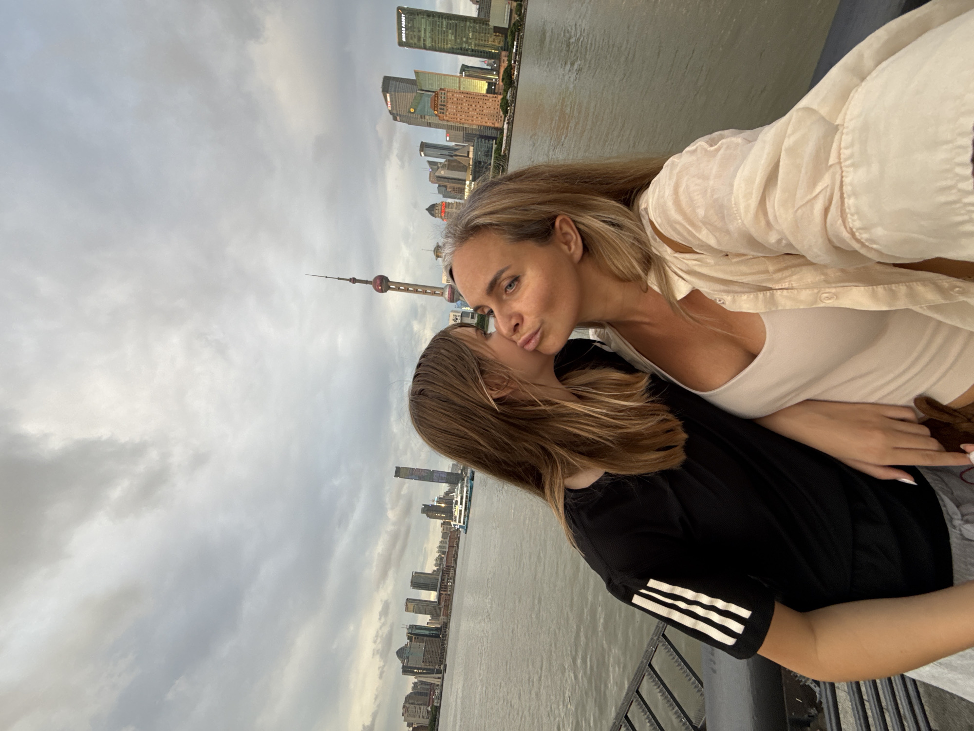 An outdoor, medium shot shows a young woman kissing a middle-aged woman on the cheek with a cityscape in the background. The young woman is on the left, wearing a black t-shirt with white stripes on the sleeve and grey pants. Her long, brown hair covers part of her face. The middle-aged woman is on the right, wearing a cream-colored shirt over a white tank top. She has blonde hair and blue eyes, and she is puckering her lips. The background features a body of water, a bridge railing, and a skyline with tall buildings, including the Oriental Pearl Tower, under a cloudy sky.