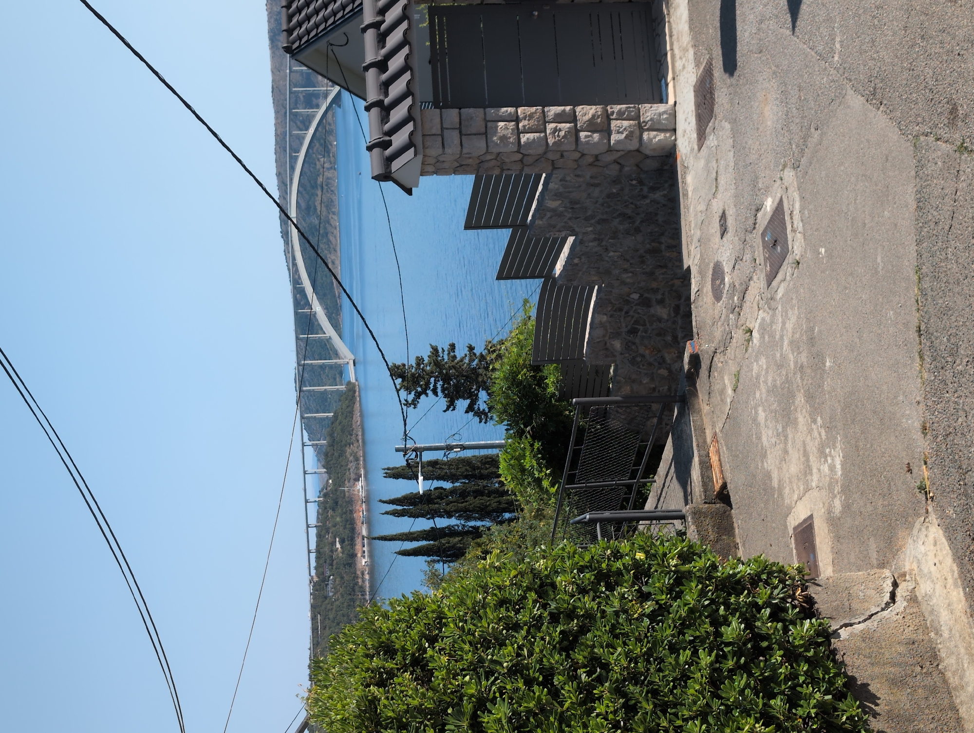 The image shows a view from a paved pathway overlooking a body of water with a large arched bridge in the distance. A lush green bush occupies the left foreground, with a stone and metal fence lining a pathway that descends into the foreground. A dark gray gate is visible on the right, constructed with stone pillars. Overhead power lines are visible against the clear blue sky. The environment suggests a sunny day in a coastal area, consistent with Kraljevica, Croatia, known for its bridges spanning across bays. No people are visible in the image.