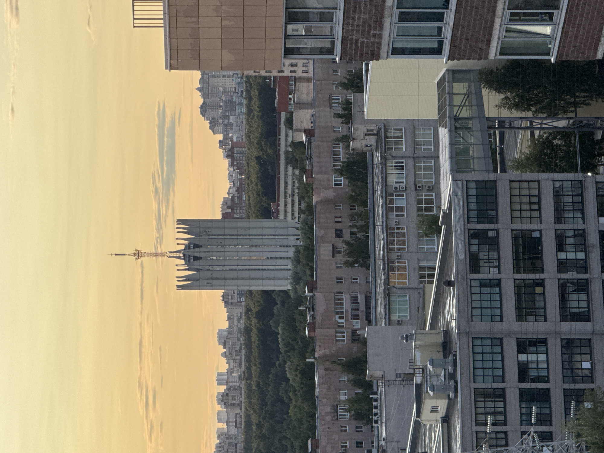 The image depicts a cityscape at dusk, with a warm, orange-tinted sky. In the foreground, a multi-story brick building with numerous windows is visible on the right side. Adjacent to it, a taller, grey concrete building with a patterned facade and a railing on its roof is partially obscured. Further into the mid-ground, a cluster of apartment buildings with varying architectural styles and brick facades are seen, interspersed with trees. In the background, a distinctive, tall concrete tower with a spired antenna stands prominently, flanked by more apartment buildings and a dense line of trees. The overall scene suggests an urban environment with a mix of residential and industrial architecture, under a soft, diffused light. The specific location is identified as Unknown, Russia.
