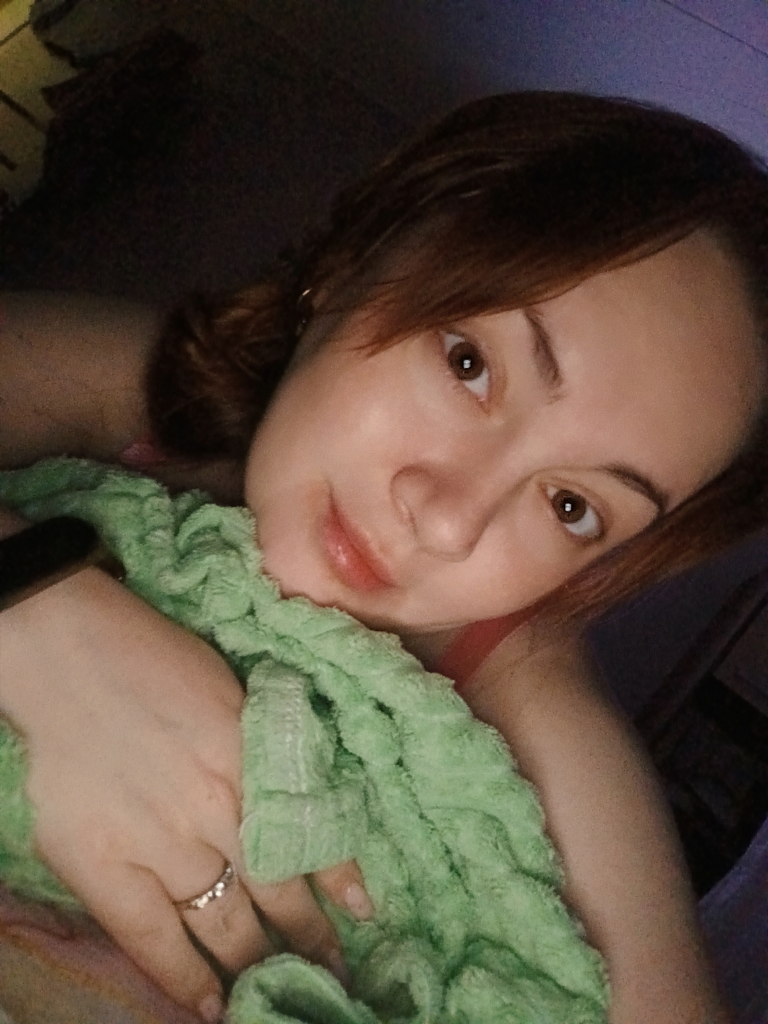 A woman is pictured in an indoor setting. She is lying down, with her head tilted slightly towards the camera. She has fair skin, brown eyes, and brown hair pulled back. She is holding a green, textured towel or cloth. She has a ring on her finger. The background appears to be a dark room, with indistinct objects. The setting and subject's state suggest a personal, informal situation. There is no information to confirm or deny that the photo was taken in Chaikovsky, Russia.