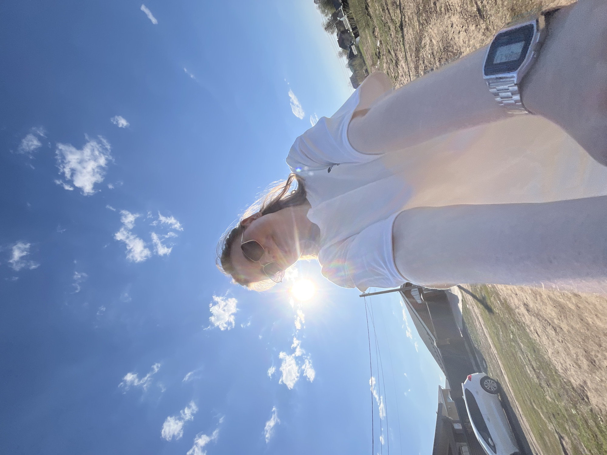 The photograph depicts a person in Chaikovsky, Russia, facing the camera with the sun in the background and wearing sunglasses and a white top. The sky is bright blue with scattered white clouds. The ground and nearby buildings are visible. The person is also wearing a digital watch. A white car is parked beside the building in the background.