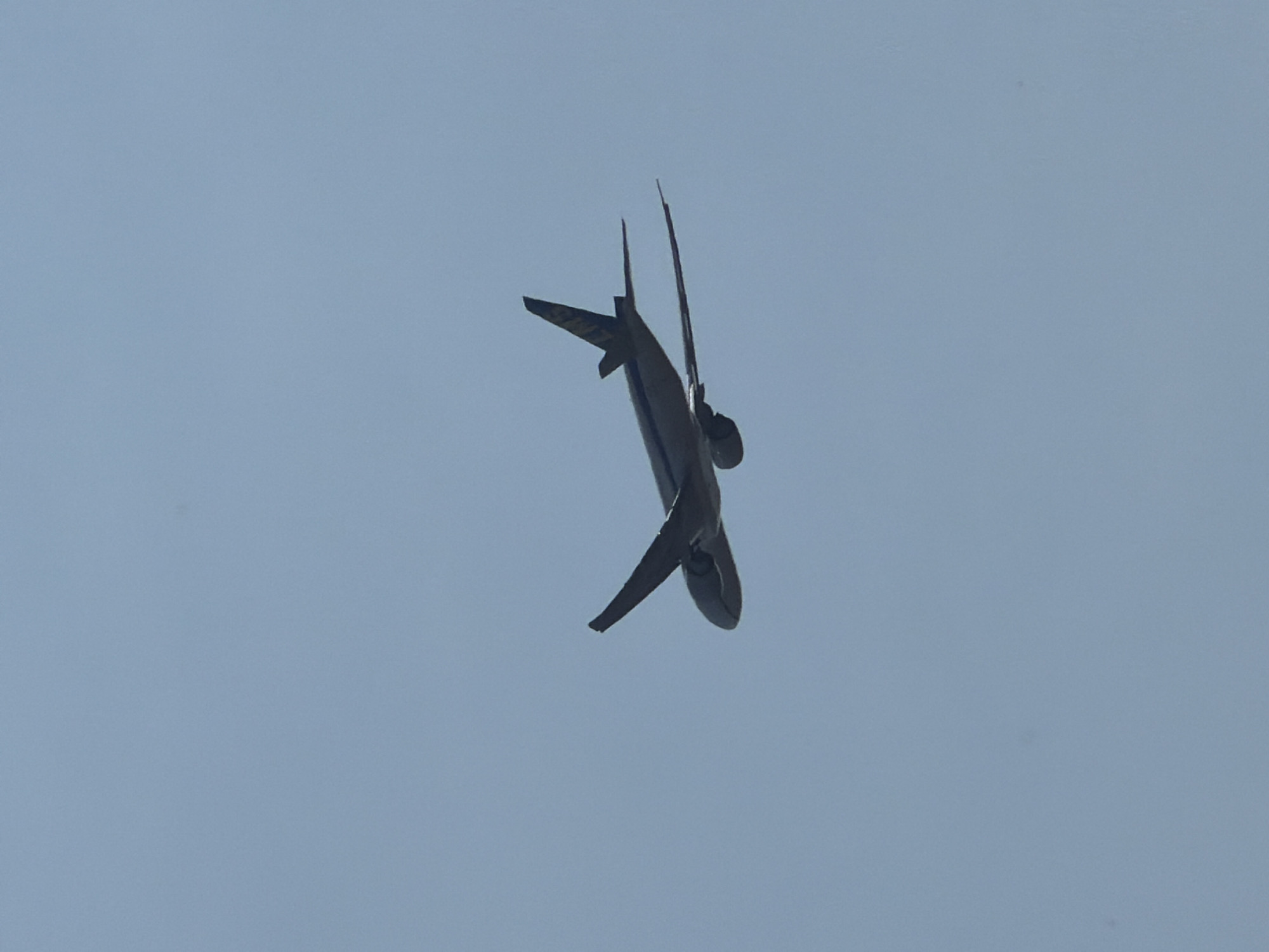 A dark-colored airplane with a yellow logo on its tail is captured in mid-flight against a clear blue sky. The airplane is angled with its nose pointing slightly upwards and to the left. Its wings are extended, and the engines are visible underneath them. The tail of the airplane has a distinctive shape and features a prominent yellow emblem. The sky is a uniform light blue, with no clouds visible. The image was taken from below the aircraft, looking up. The location is the city of Niederanven, Luxembourg.