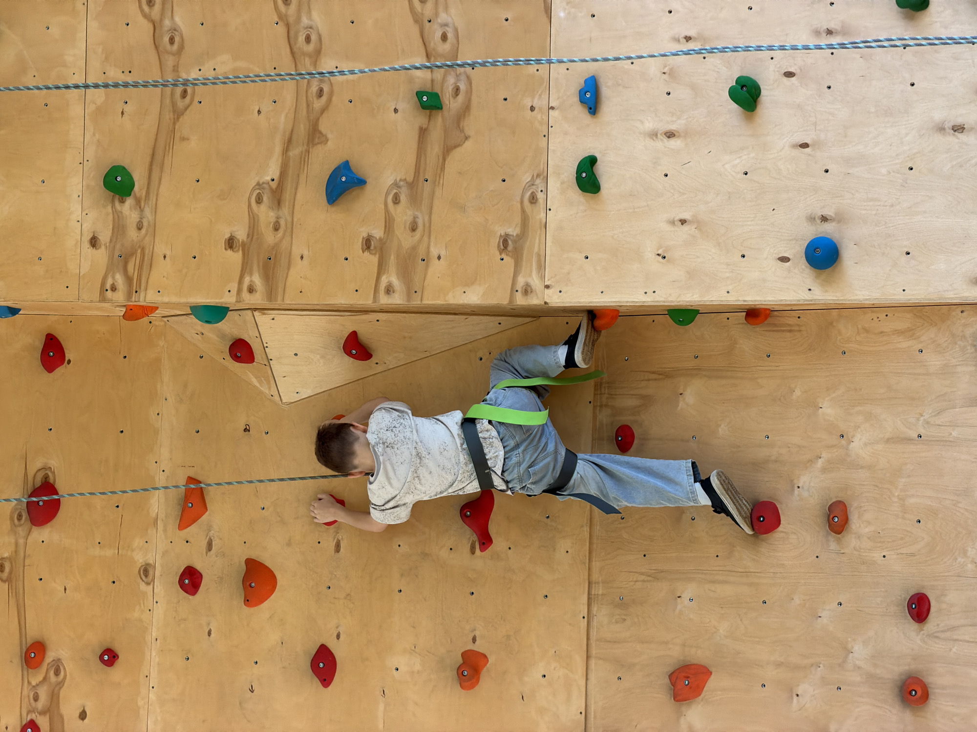 A young person is climbing an indoor rock wall. They are wearing a climbing harness and jeans. The climbing wall is made of wood and has various colored climbing holds attached to it. A rope runs across the top of the wall. The location is not specified in the image and cannot be confirmed as Izhevsk, Russia.