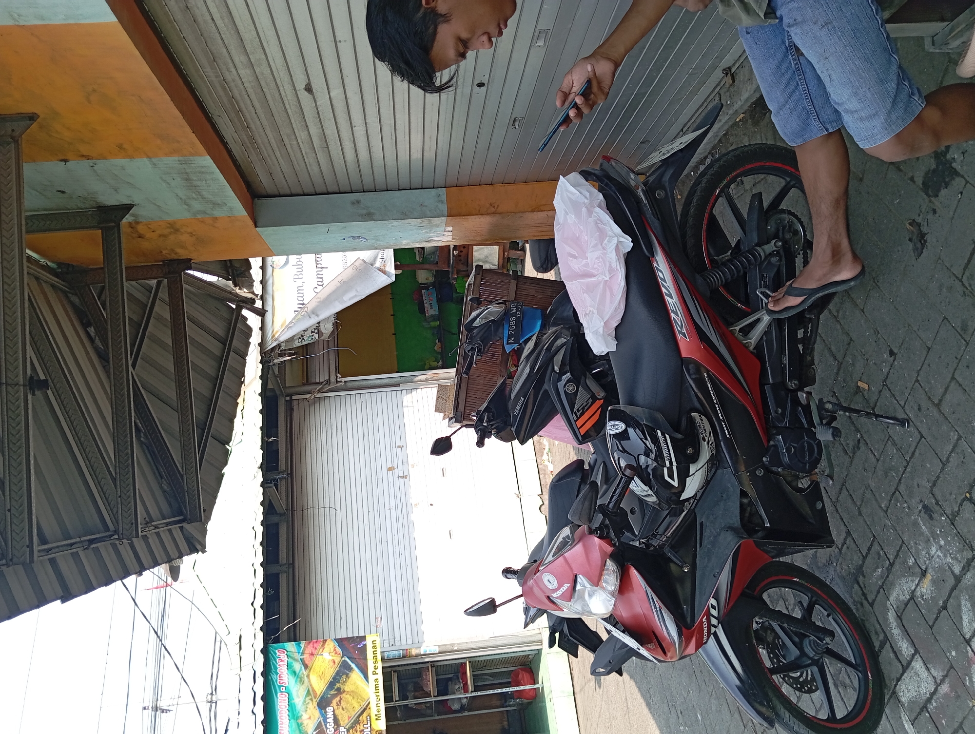 A man, identified by his presence in the image, sits on a pavement, looking at a mobile phone. He is wearing denim shorts and a t-shirt. Next to him are two parked motorbikes, one red and black and the other black. A white plastic bag is on the seat of the red and black motorbike. The scene appears to be in front of a shop or building with a corrugated metal shutter and an orange and light blue painted wall. A banner advertising food is visible on the left side of the image. The area looks like a street scene in Kepulungan, Indonesia, based on the context provided. The pavement is made of interlocking bricks. The sky is overcast. The overall impression is a candid street scene in Indonesia, possibly in an urban or semi-urban commercial area.