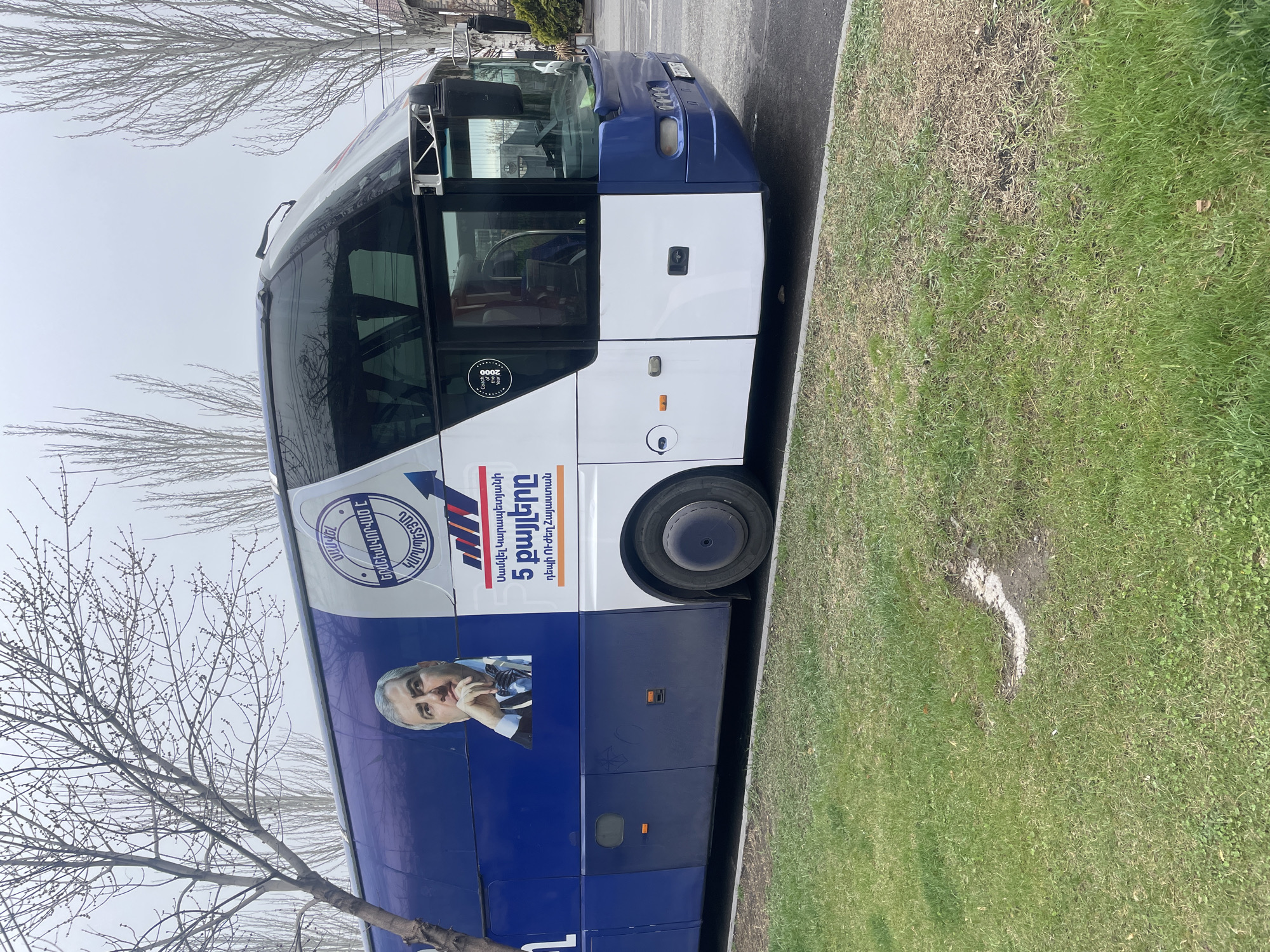 A large commercial coach bus, painted in a two-tone royal blue and white livery, is parked parallel to a paved road in Yerevan, Armenia. The side of the bus displays a prominent vinyl wrap featuring a portrait of a man with dark hair, grey at the temples, wearing a dark suit and bow tie, with his right hand resting under his chin. Accompanying Armenian text reads, "Samvel Karapetyan's 5 steps towards a strong Armenia," and another circular logo with Armenian text including "SMAGAN." A small "Coach of the Year 2000" sticker is visible on the passenger door area. The bus's front section is blue, transitioning to white, with visible headlights, grille, and windshield wipers. The vehicle's tires are black, with a silver hubcap visible on the rear wheel. To the left of the bus, a thin, bare-branched tree stands on a patch of green grass with some dry, brown areas. In the background, numerous other bare deciduous trees line what appears to be a street or parkway. The sky is overcast and grey. No individuals are visible in the scene.