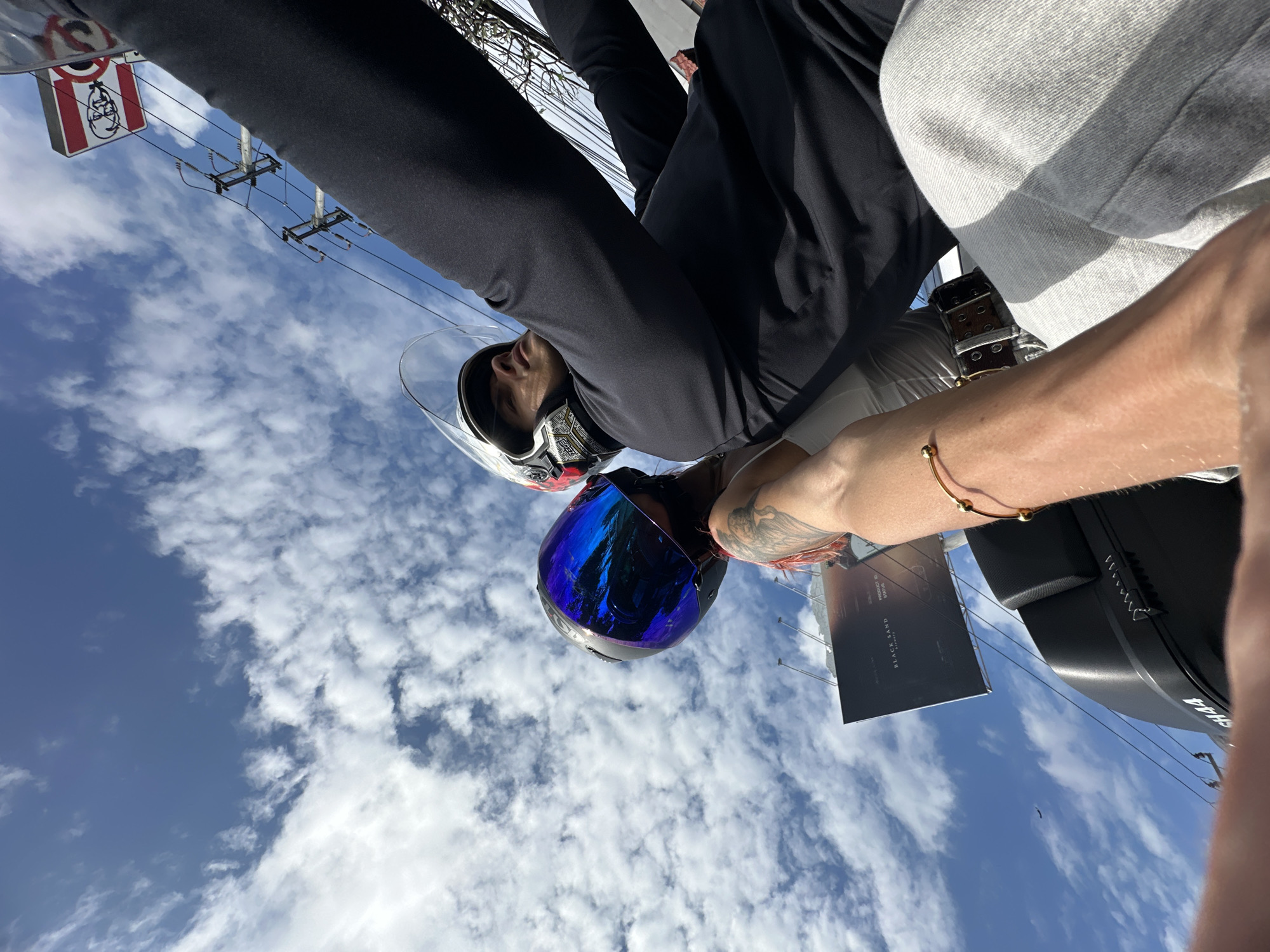 A low-angle shot showing two individuals on a motorcycle in Kuta Selatan, Indonesia. The view is from the perspective of the person on the back of the motorcycle and oriented towards the sky filled with scattered clouds. A person in a white and black helmet is in the front, and another person in a blue tinted helmet is behind the rider. The person in the back is wearing a black long-sleeve shirt and pants. The person taking the photo is wearing a light gray shirt and a leather belt. They have a gold bracelet on their wrist and a tattoo on their arm. They are holding a black rectangular object. In the background, a sign for KFC and electric power lines are visible. A black object is visible to the right.